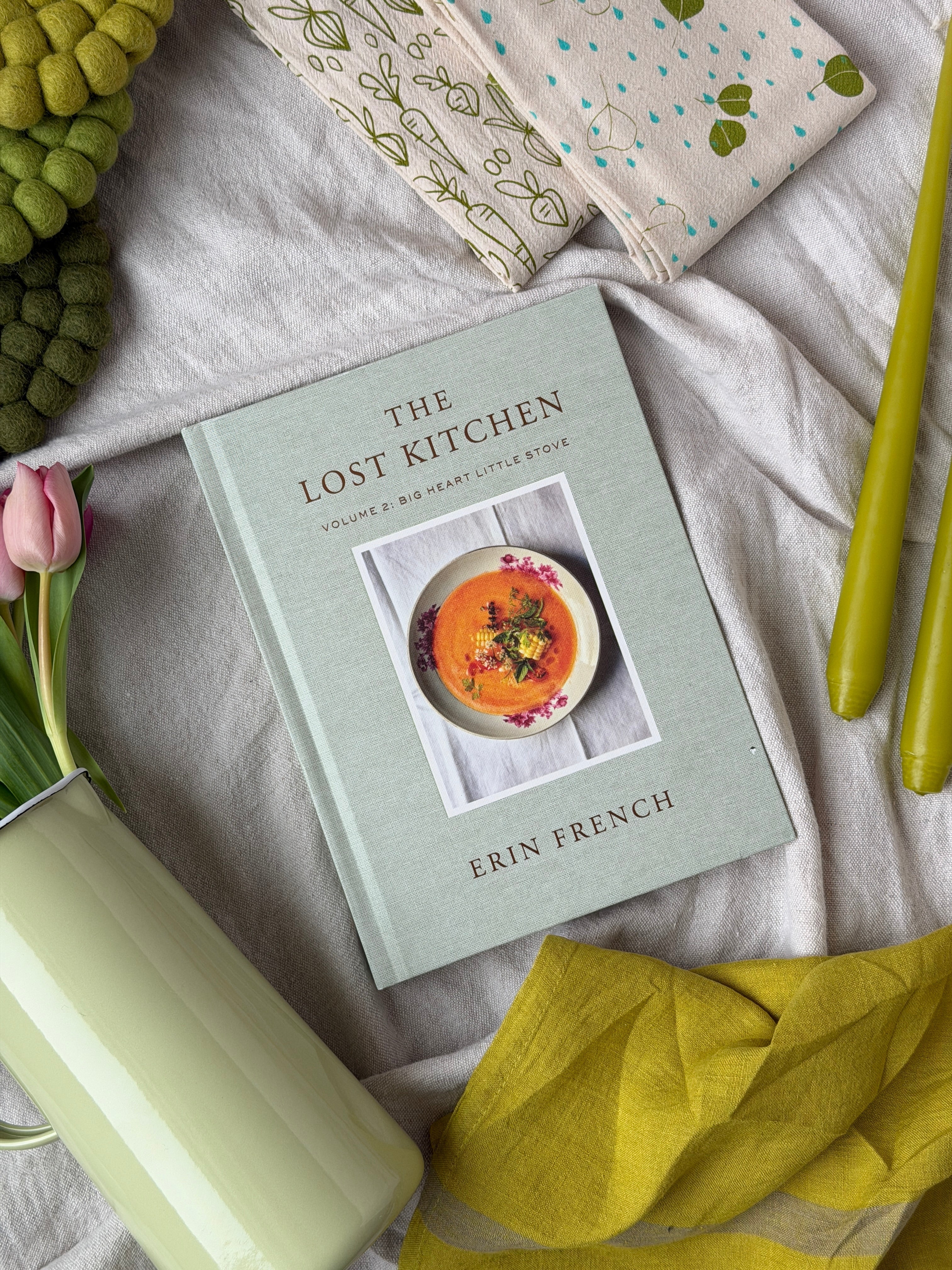a spring flatlay with a ceramic pitcher filled with light pink tulips, green tea towels, green tapered candles, and the lost kitchen cookbook in the centre.