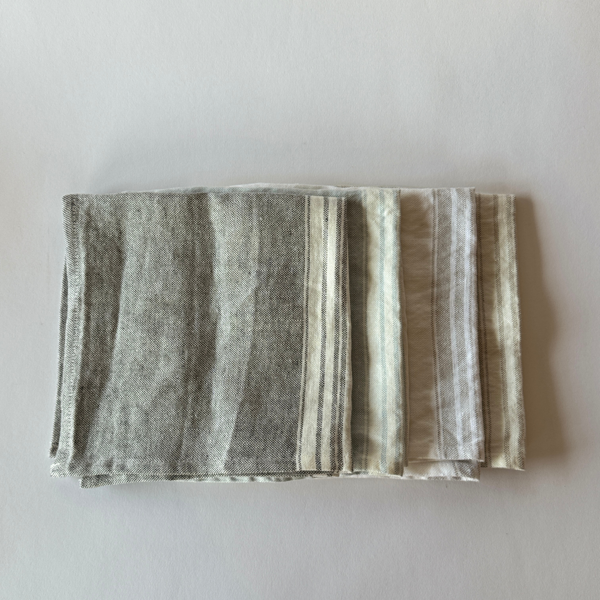 Three Linen Way - Maison Table Napkins by Grace & Company, in gray and white, stacked on a light surface.