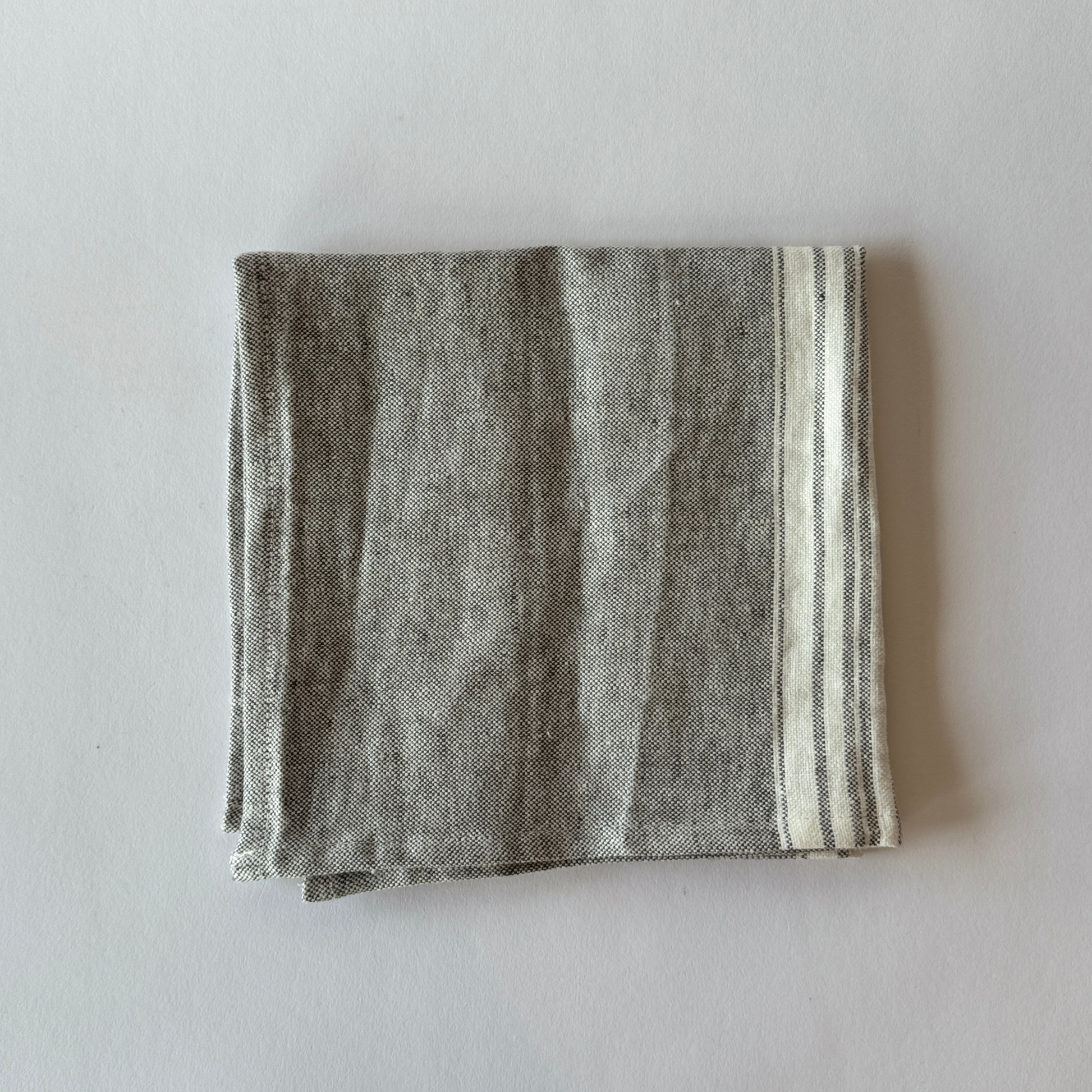 The Grace & Company Linen Way - Maison Table Napkin is folded, gray with a white striped edge, on a light surface.