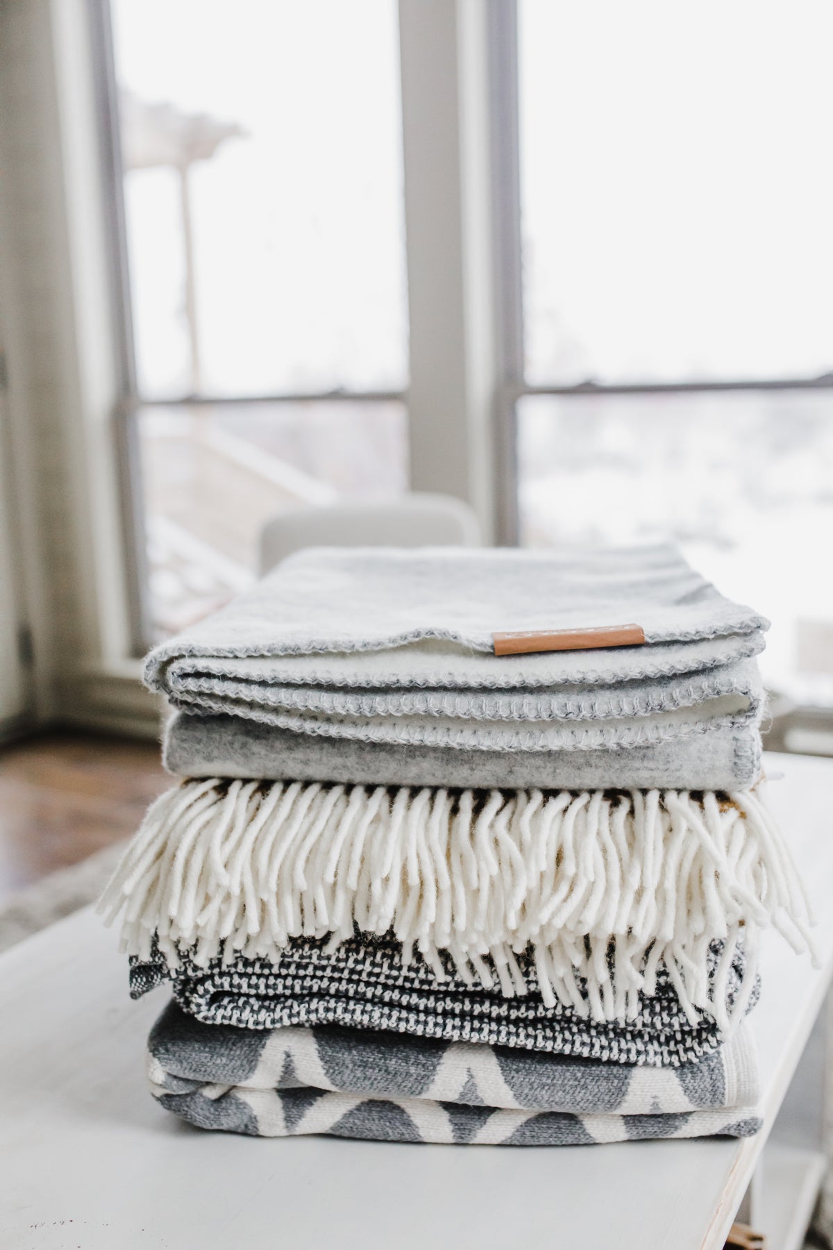 A stack of folded, cozy blankets in neutral colors sits on a white table by large windows.
