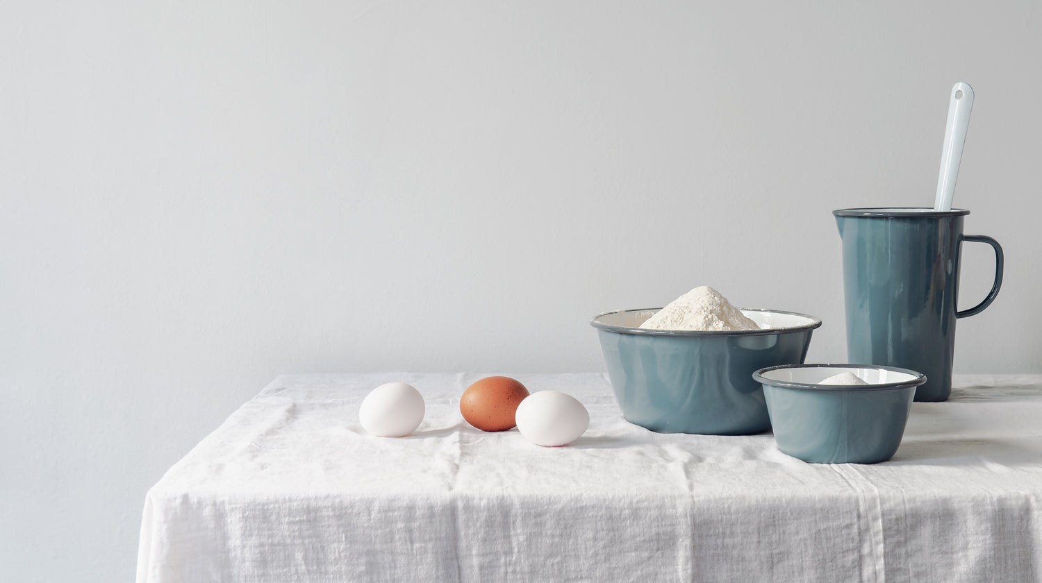 Three eggs, a bowl of flour, a blue mug, and a blue bowl on a white table covered with a white cloth.