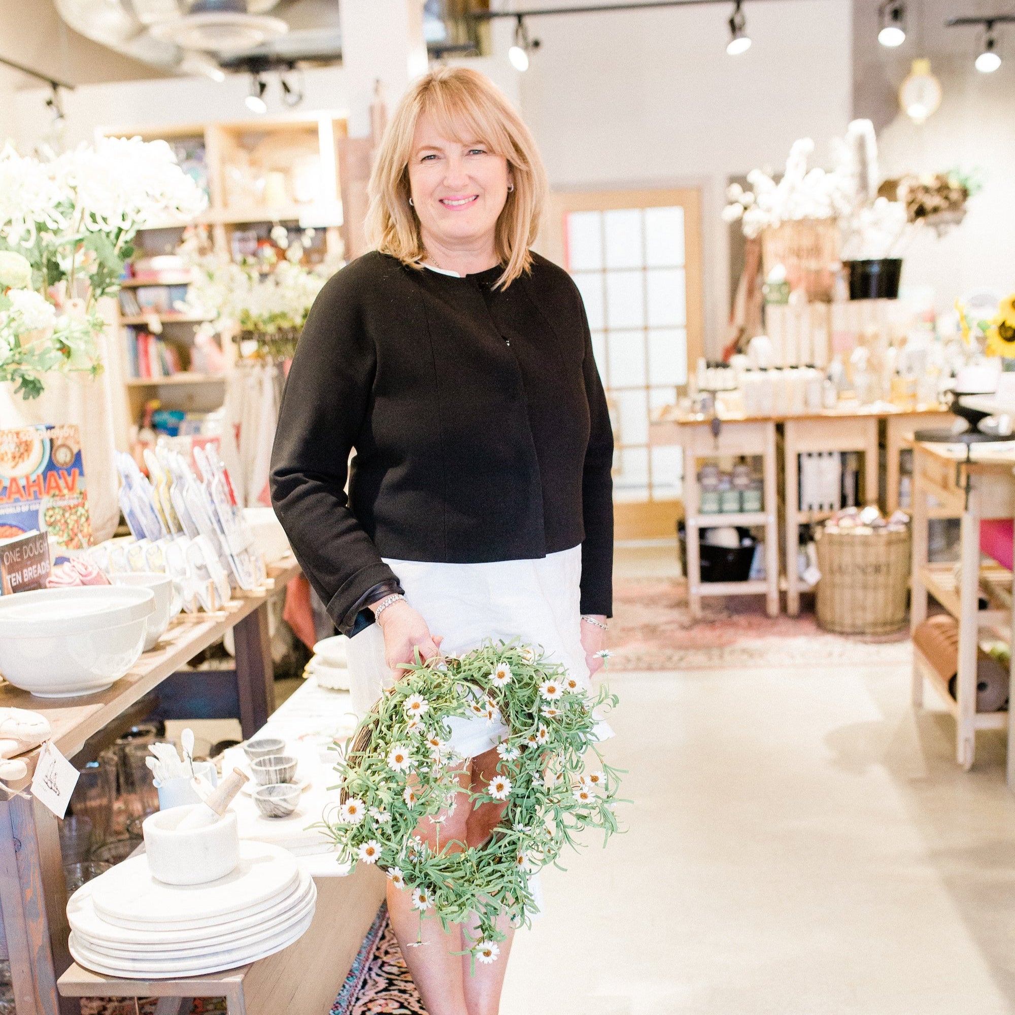 Smiling woman in a shop holding a floral wreath, surrounded by home decor items and shelves.