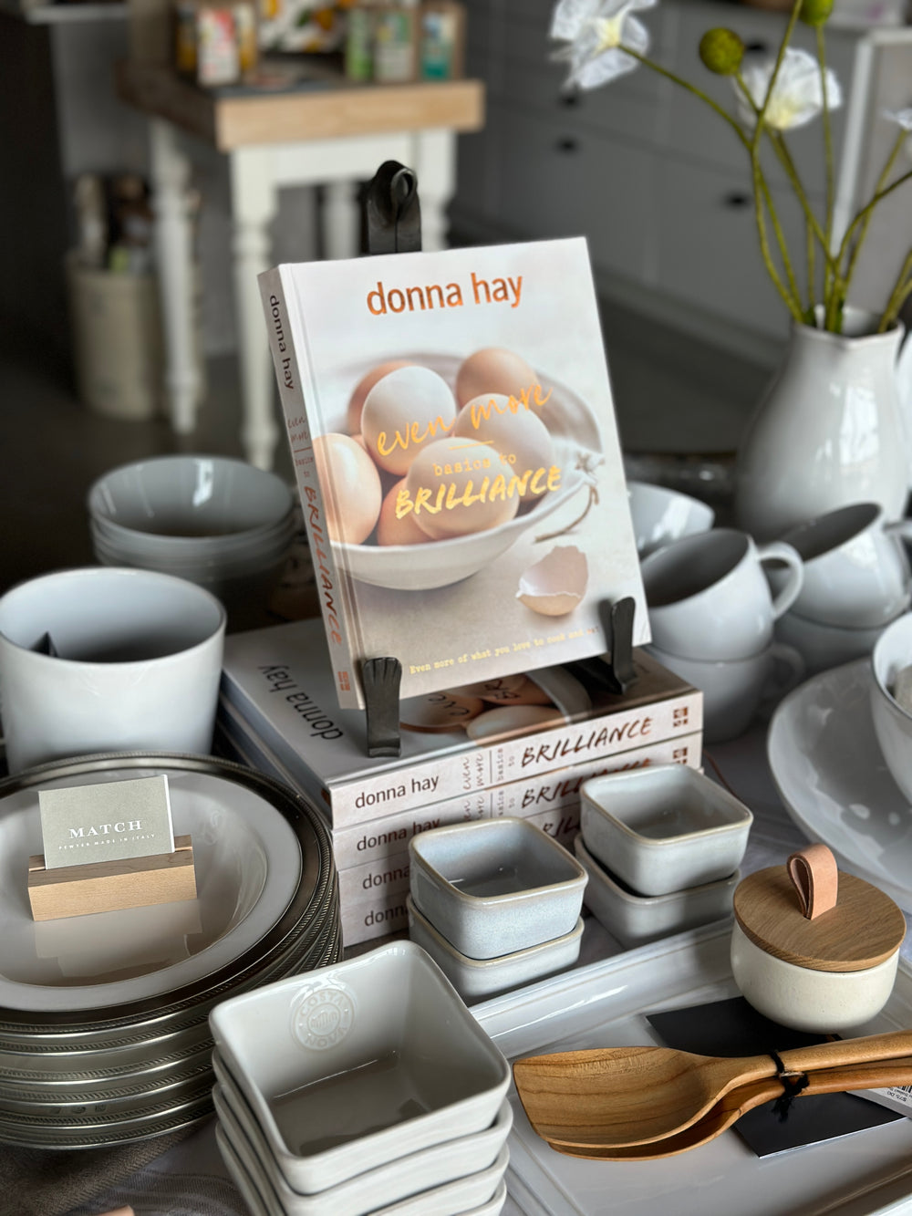 Cookbook on a stand surrounded by ceramic dishes, kitchenware, and stacked plates on a kitchen counter.