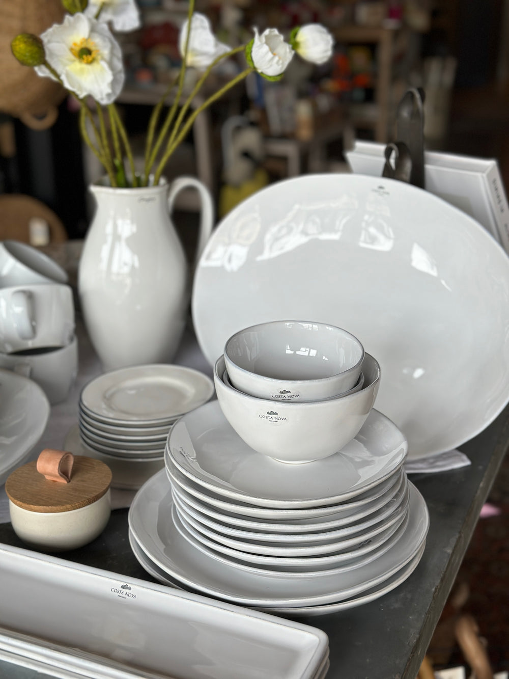 White ceramic dishes, bowls, plates, and a pitcher are arranged on a table with a vase of white flowers.