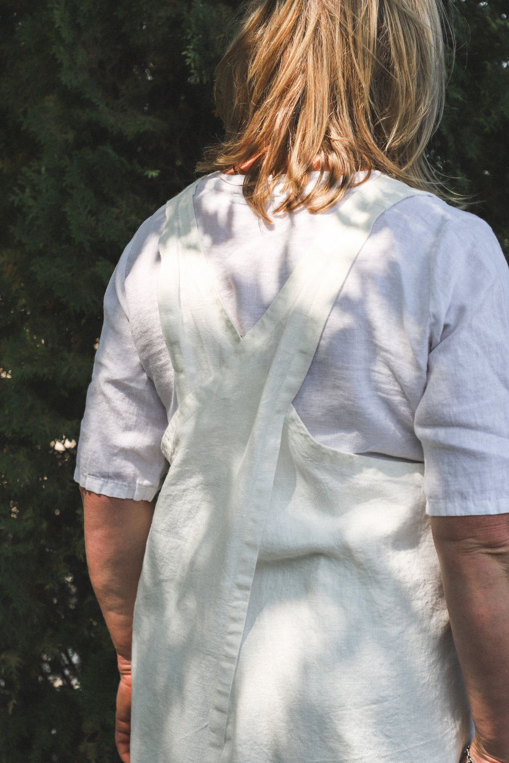 A person with shoulder-length blonde hair wears a white cross-back apron over a white shirt, standing outdoors.