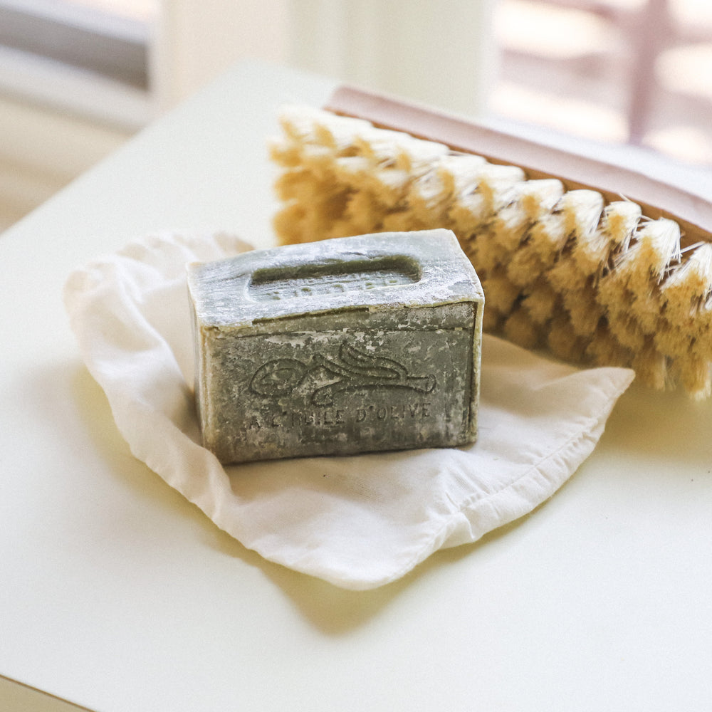 A worn bar of soap on a cloth sits beside a wooden bath brush with natural bristles on a white surface.