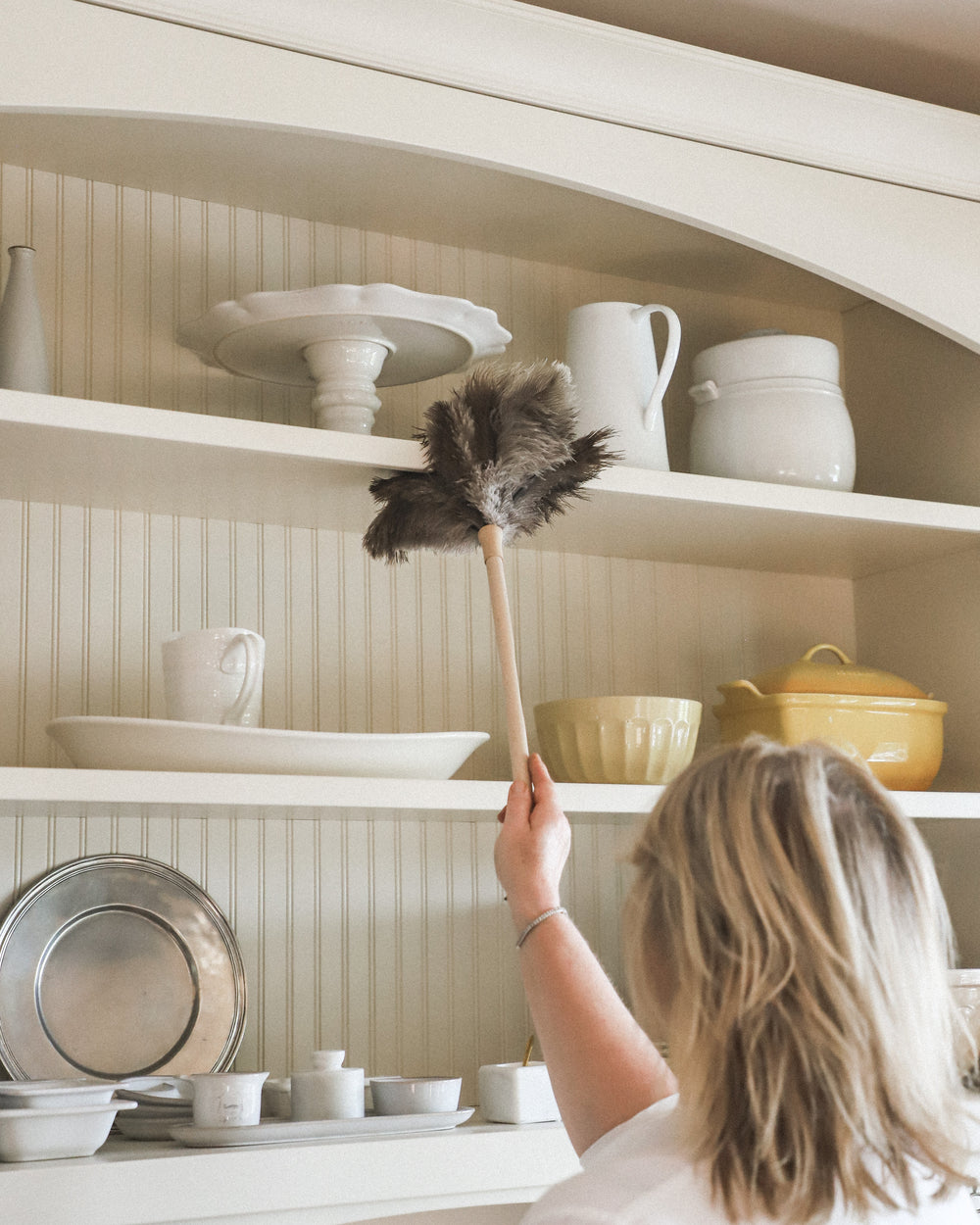 A person dusts white dishes on a high kitchen shelf using a feather duster.