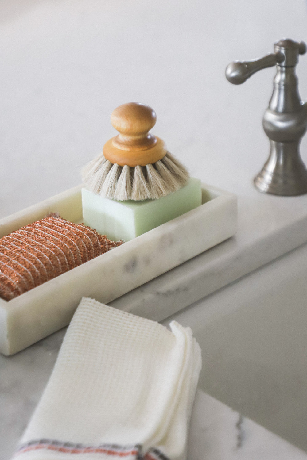 Marble tray with a scrub brush, green soap, and an orange sponge next to a sink and white towel.