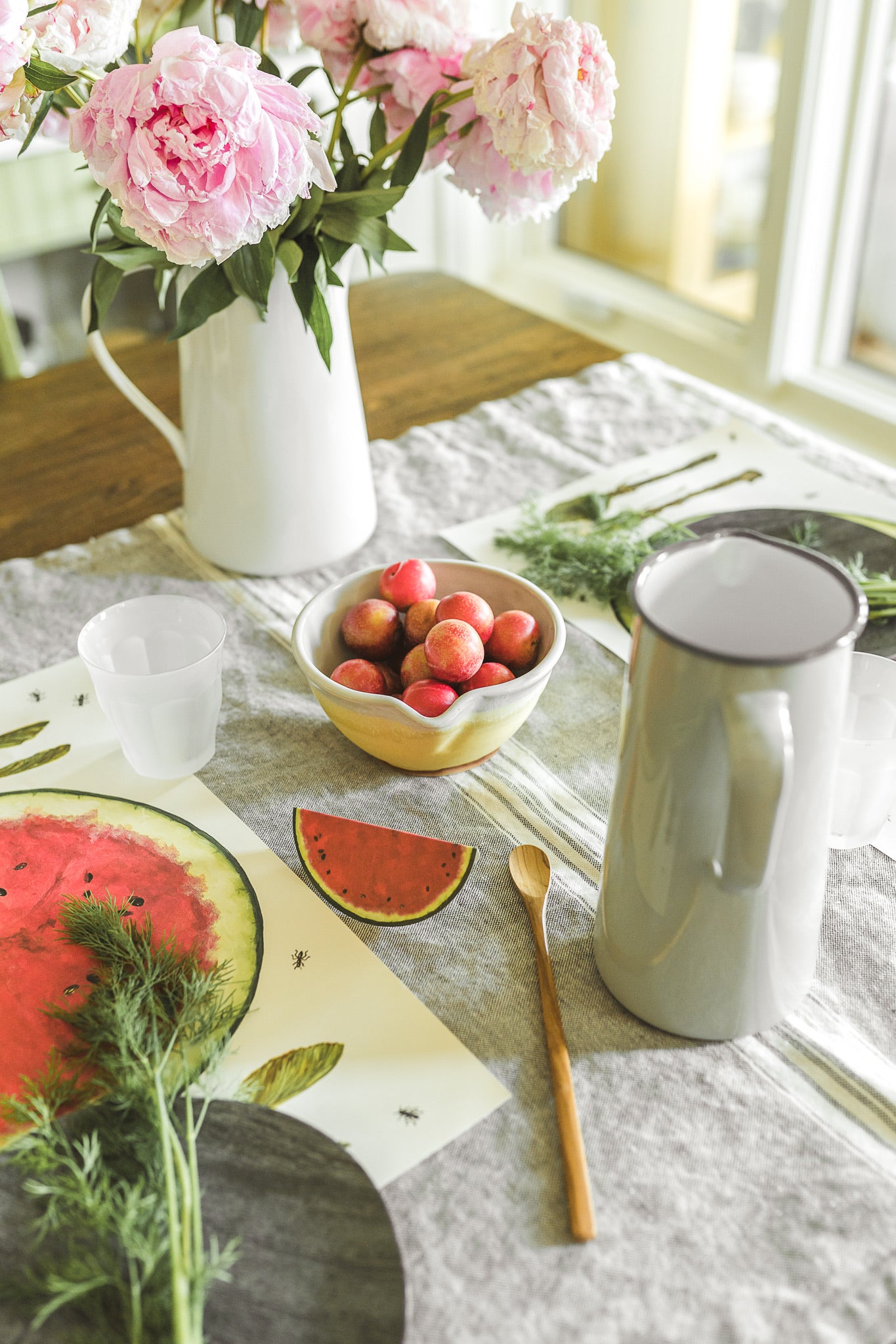 A summer table setting with pink flowers, a bowl of plums, a watermelon placemat, and a gray pitcher.