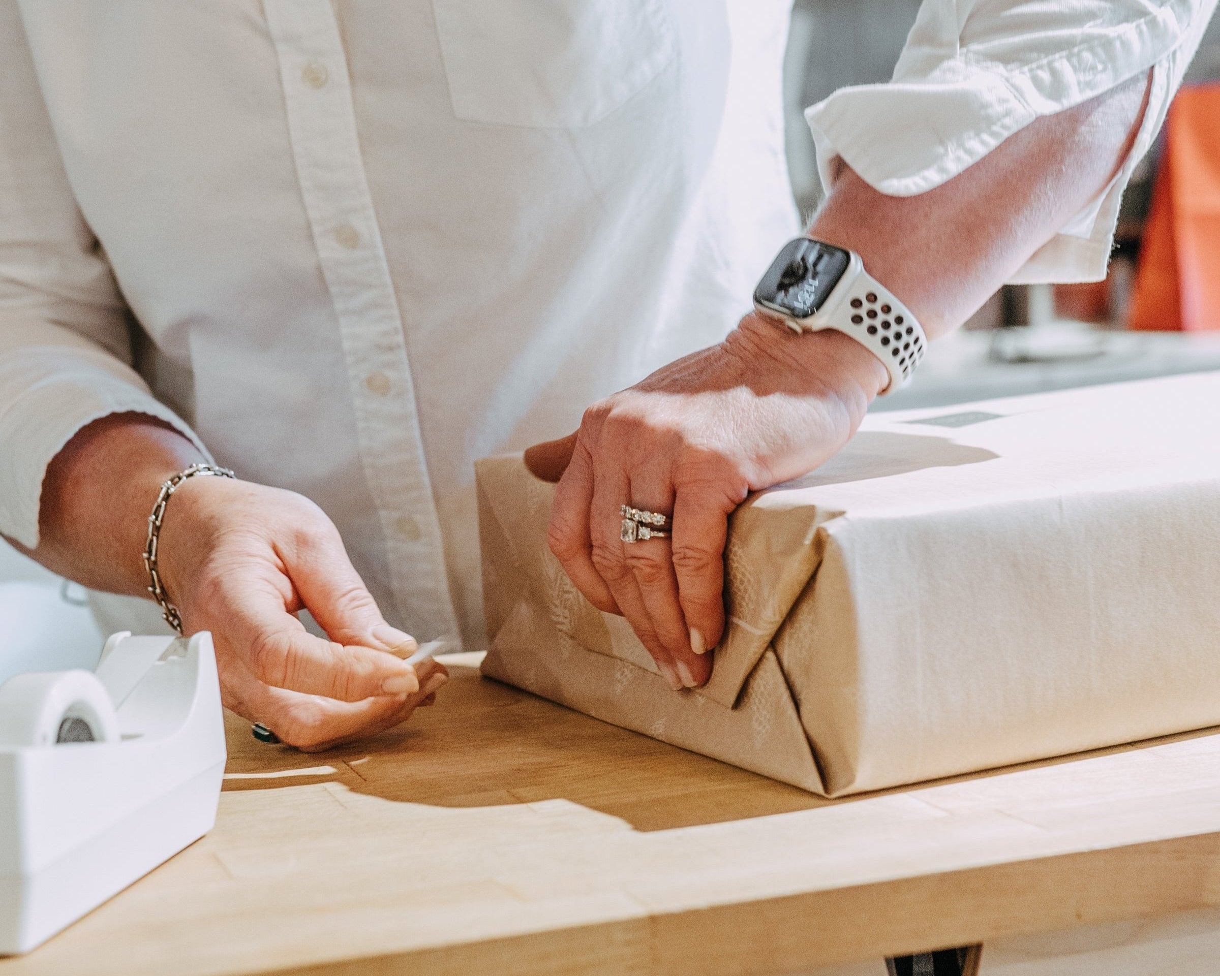 Person in a white shirt wrapping a box with brown paper on a wooden table, wearing a smartwatch and rings.