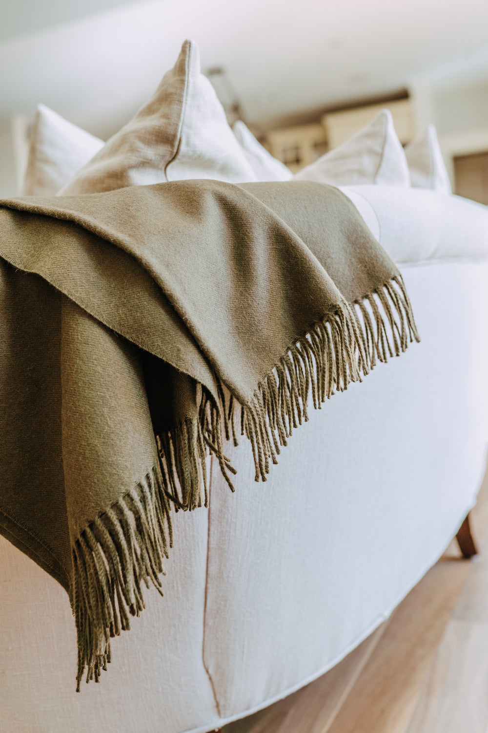 A beige sofa with cream pillows and a folded olive green fringed throw draped over the back.