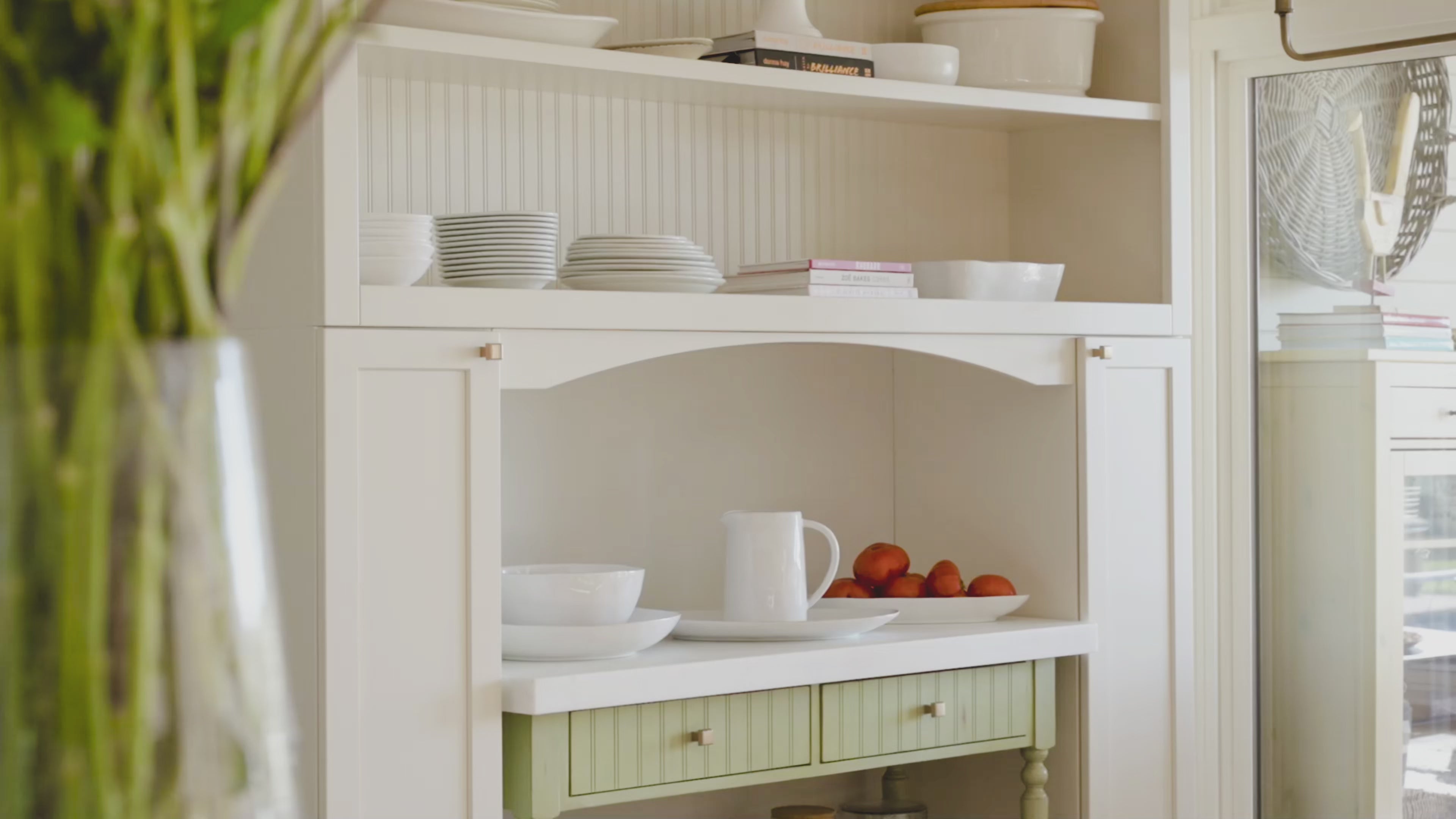 Load video: White kitchen hutch with dishes, a pitcher, books, and a bowl of oranges; flowers blurred in the foreground.