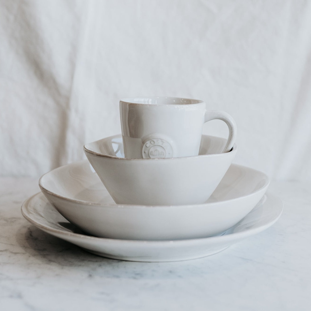 Costa Nova Nova Dinner Collection by Grace & Company stacked on marble with white fabric backdrop.