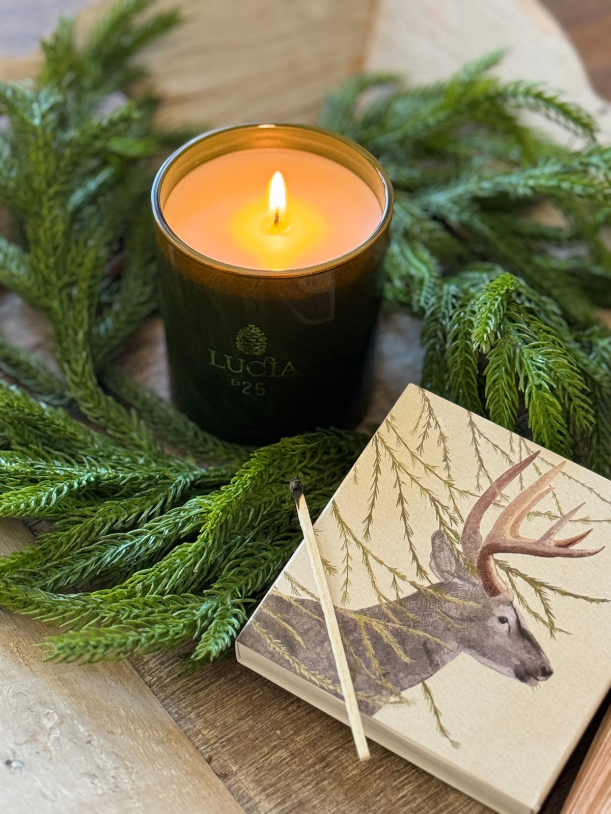 A lit green candle with greenery, next to a notepad with a deer illustration and a matchstick on a wooden surface.