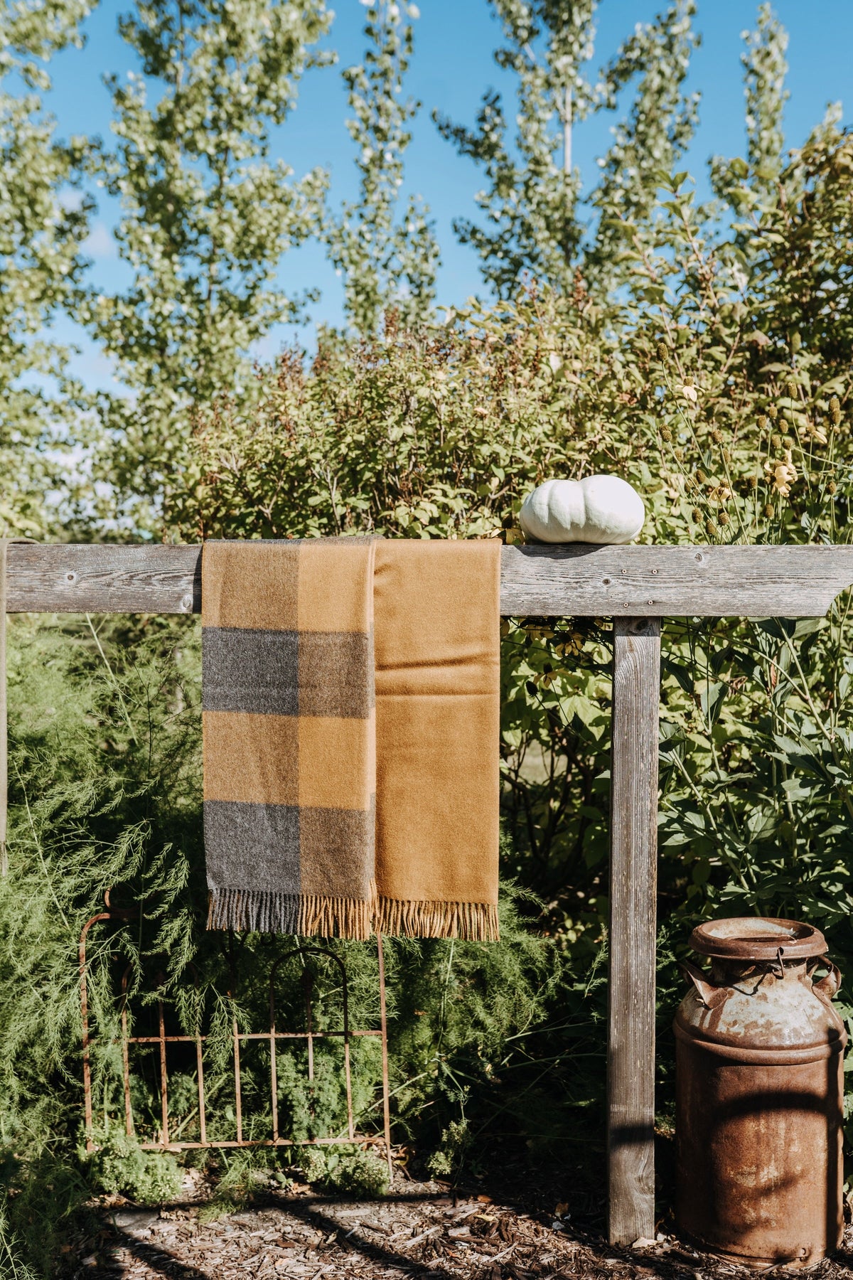A brown plaid blanket hangs on a wooden fence in a garden, with a white pumpkin and milk can nearby.