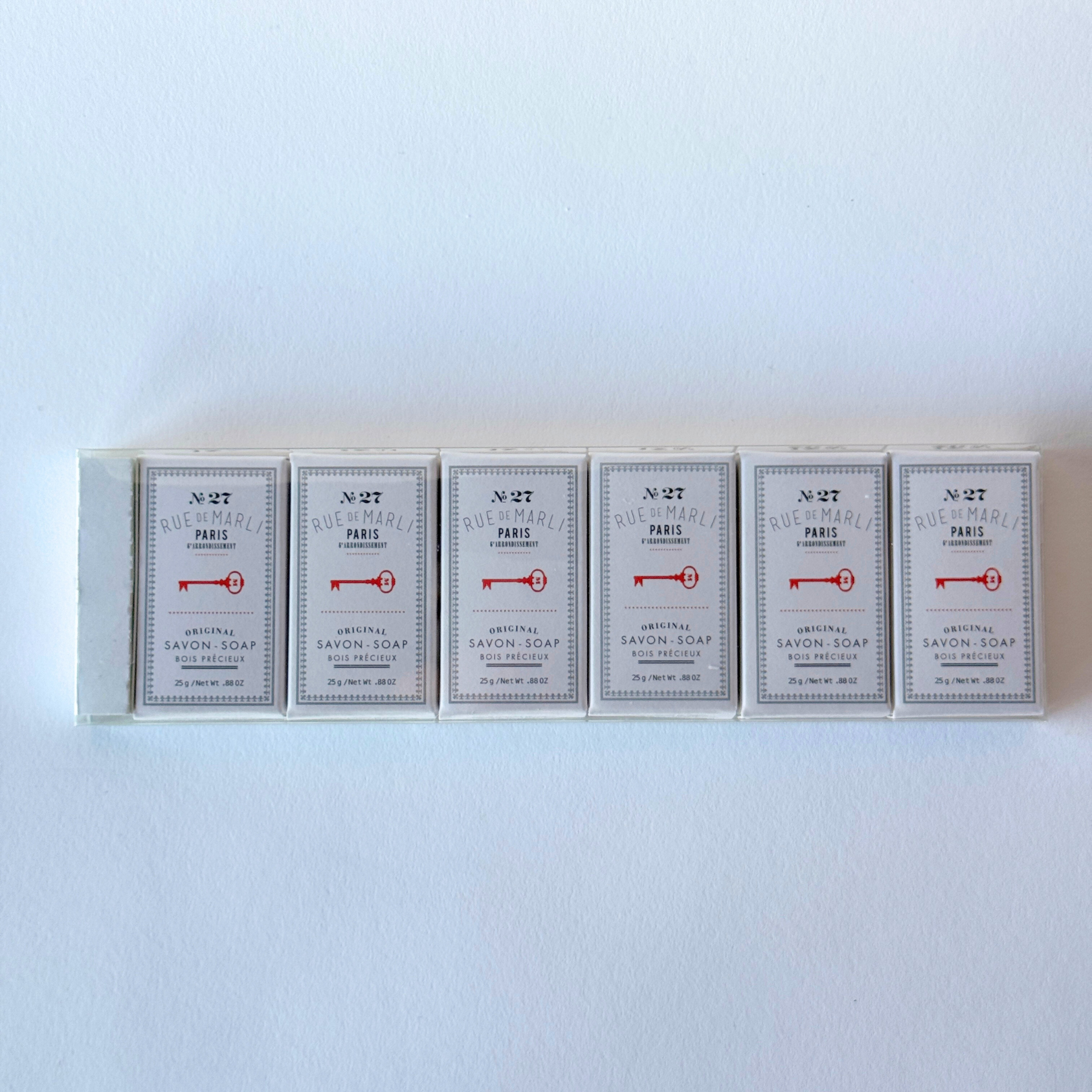 Six Rue de Marli No. 27 mini guest soaps by Grace & Company, each with white label and red keys, in a row.