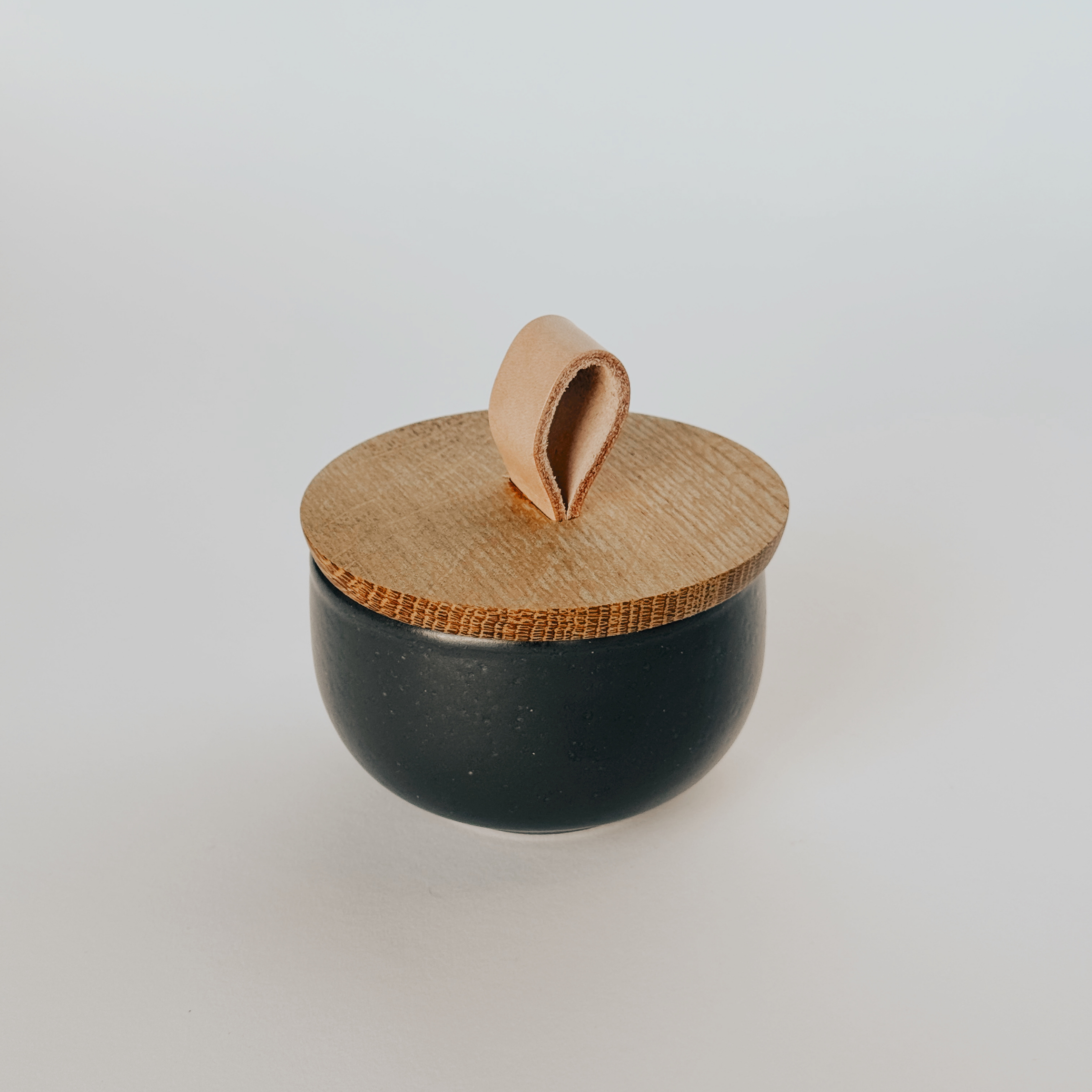 The Casafina Pacifica Salt Cellar by Grace & Company features a black container, oak lid, and looped leather handle.