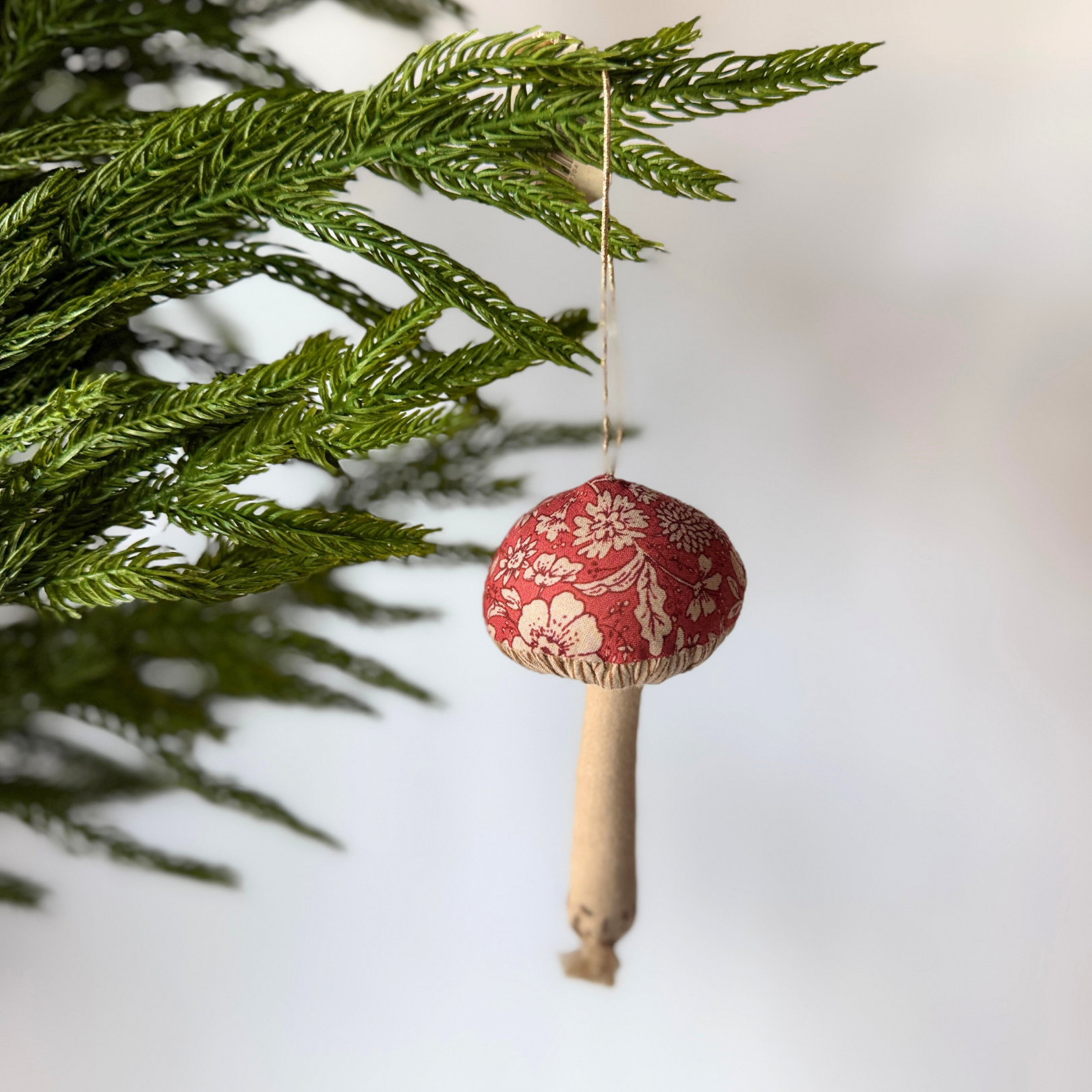 The Grace & Company Maileg - Mushroom Ornament with a red cap hangs from a green tree branch.