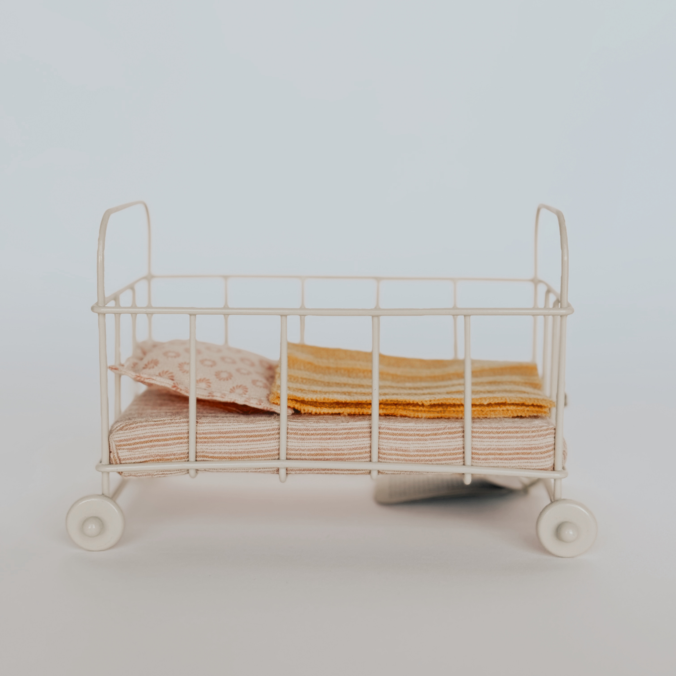 The Maileg - Baby Crib by Grace and Company Ltd is a small white toy crib with striped bedding and a patterned pillow.