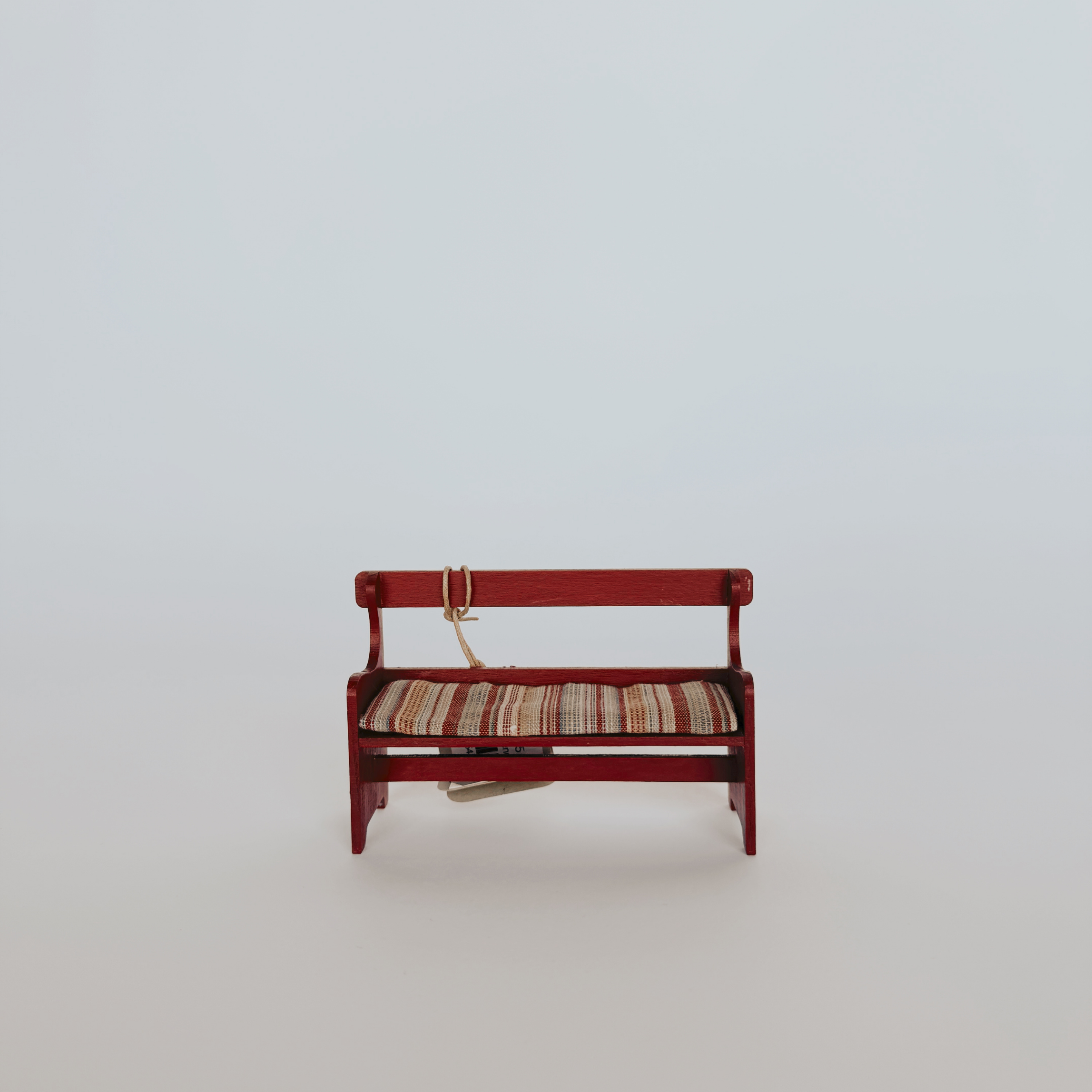 The Grace & Company Maileg - Bench with Pad is a small red wooden bench with striped cushion and strap on a plain background.