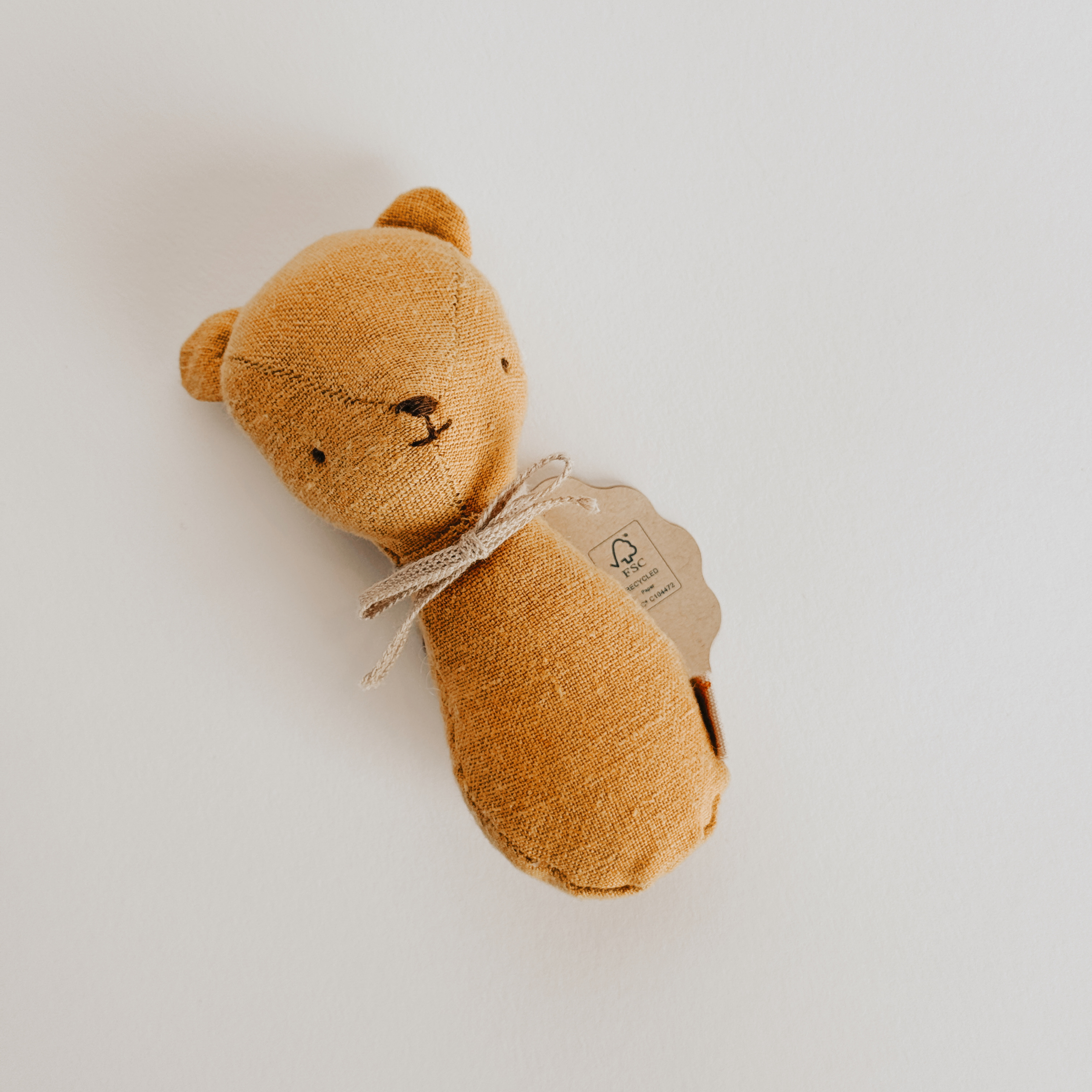 A soft brown Maileg Teddy Rattle by Grace & Company with a simple face and bow, lying on a white background.
