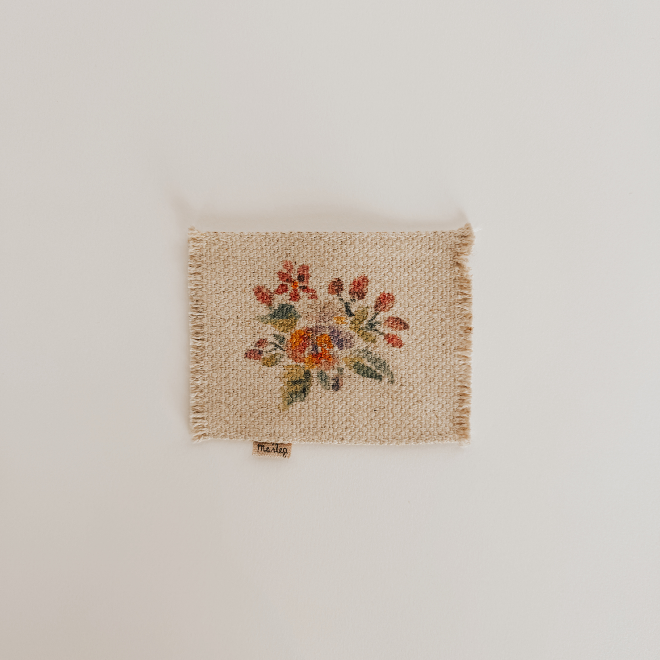 The Maileg - Stencill Carpet by Grace & Company features a colorful embroidered flower and hangs on a white wall.