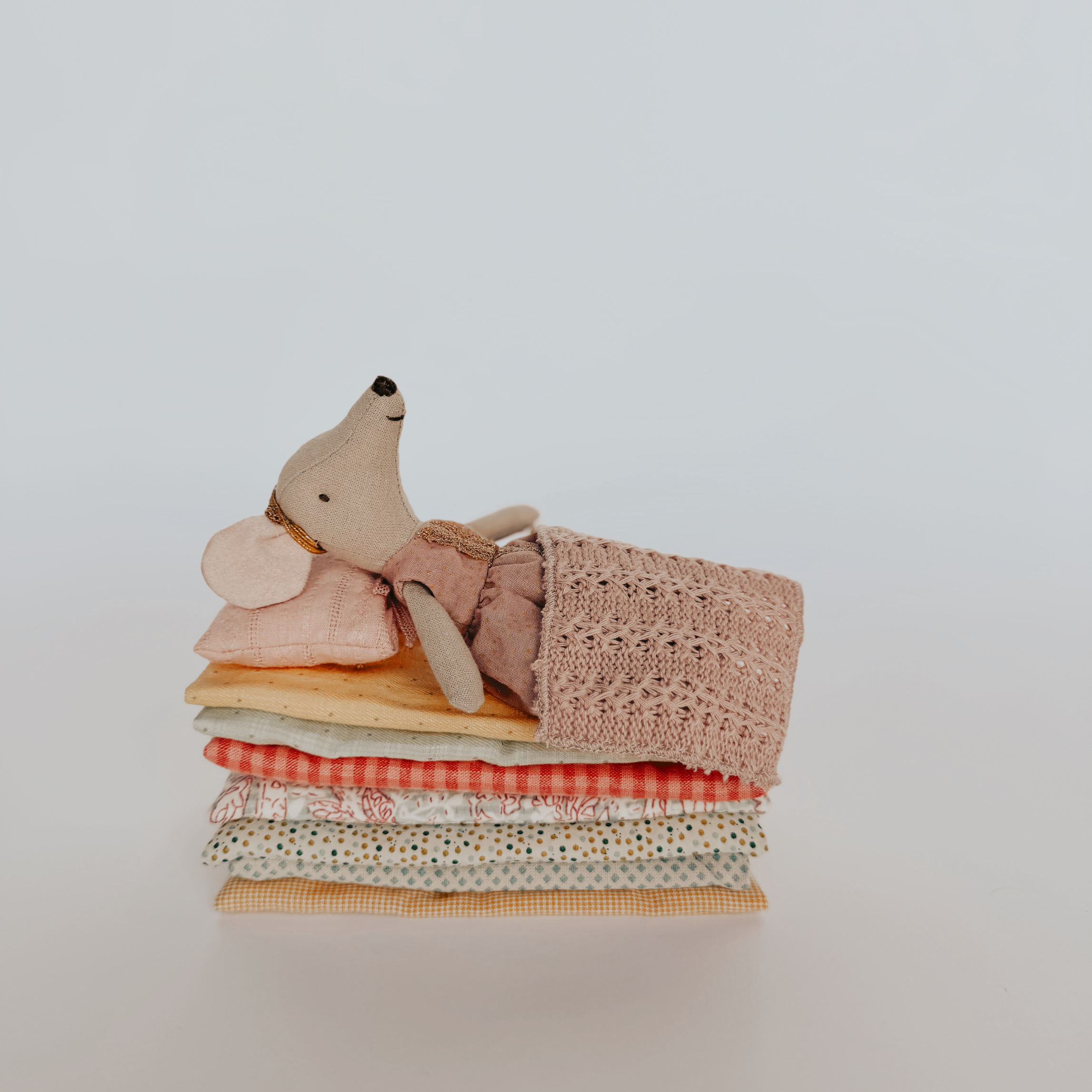 The Maileg Princess Mouse Big Sister by Grace & Company rests on stacked fabric, covered with a knit blanket.