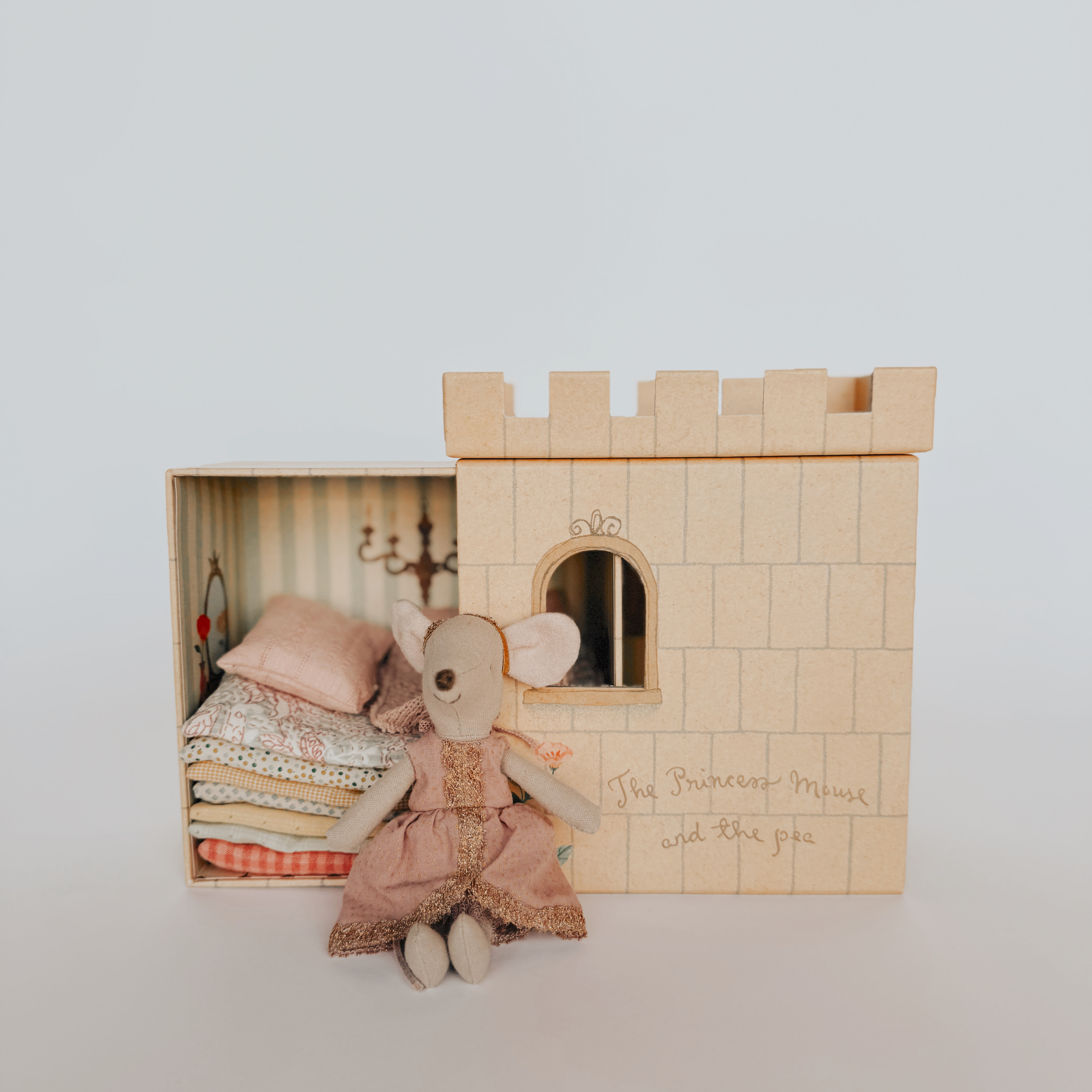 The Maileg Princess Mouse Princess and the Pea by Grace & Company sits by her toy castle and box of stacked mattresses.