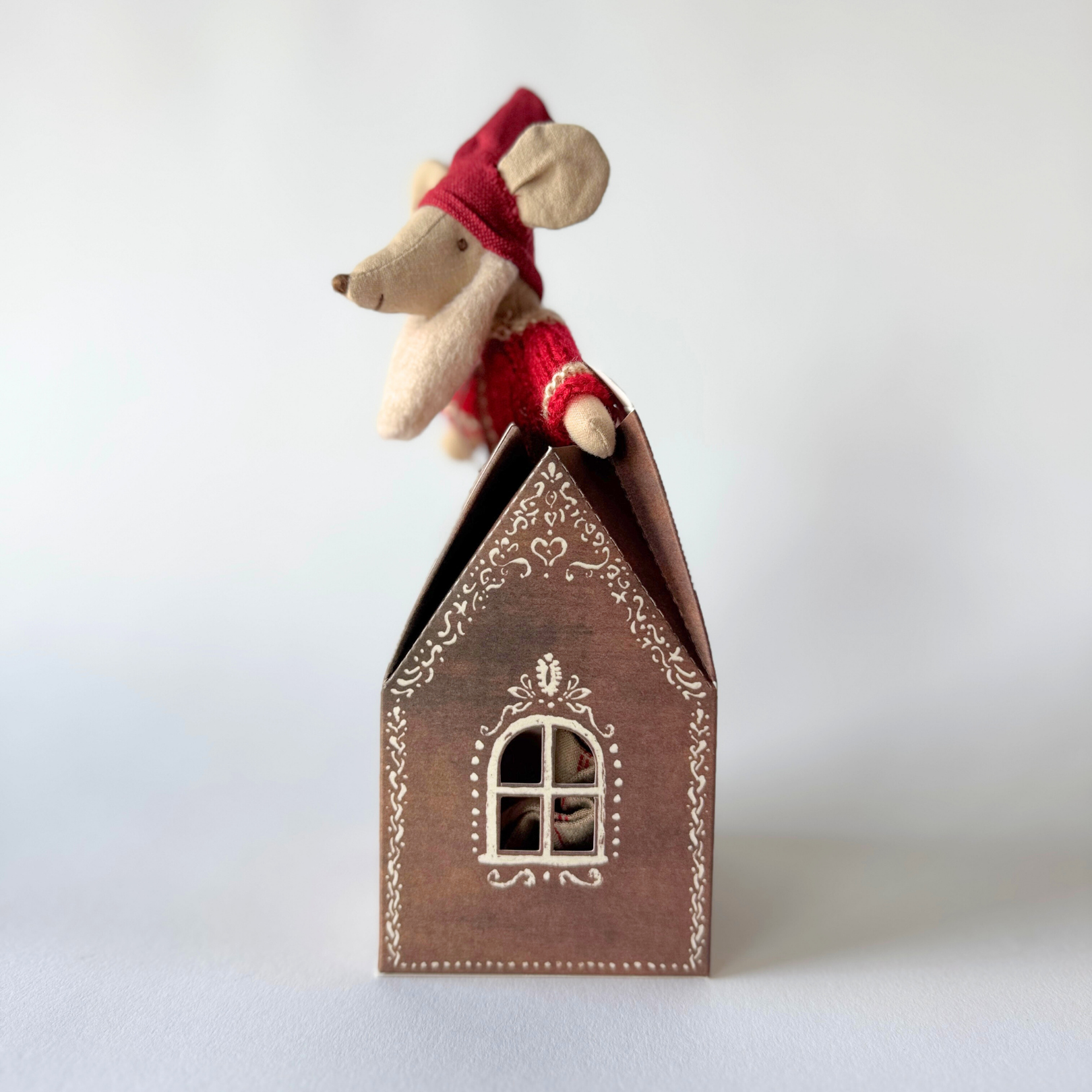 The Maileg - Santa Mouse by Grace and Company Ltd sits on a brown house with a white ornate window and patterns.