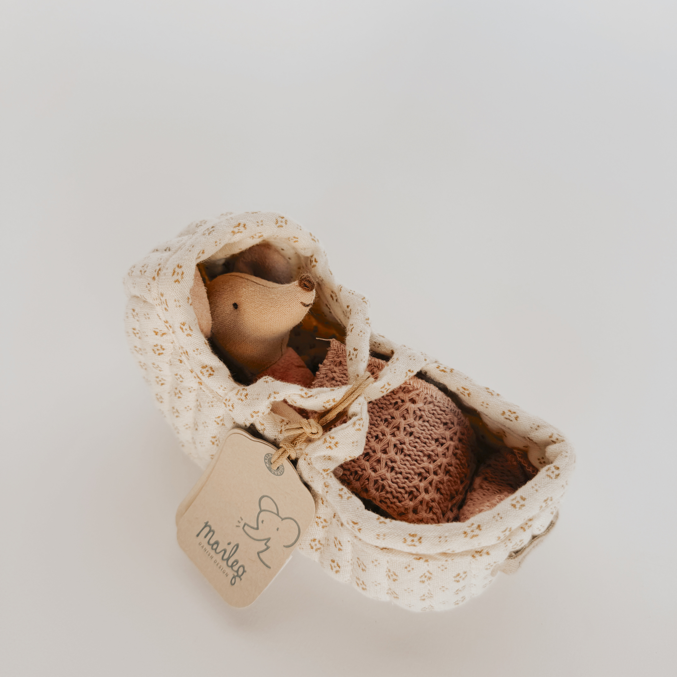 A Maileg - Carry Cot + Sister Mouse by Grace & Company ltd rests in a patterned fabric baby bassinet.