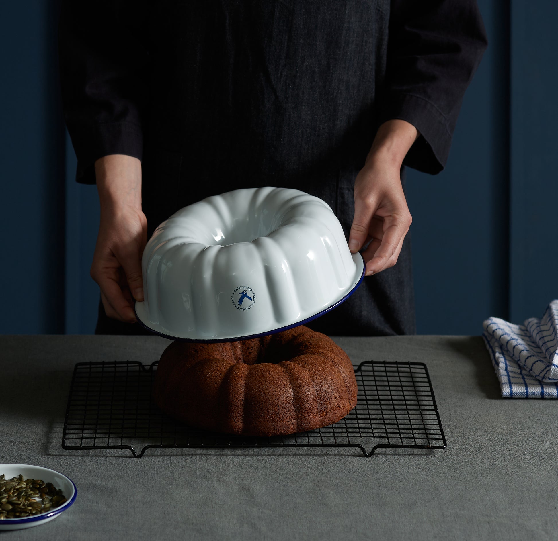 Someone lifts a Falcon Enamelware - Bunt Pan by Grace & Company off a chocolate cake on a cooling rack.