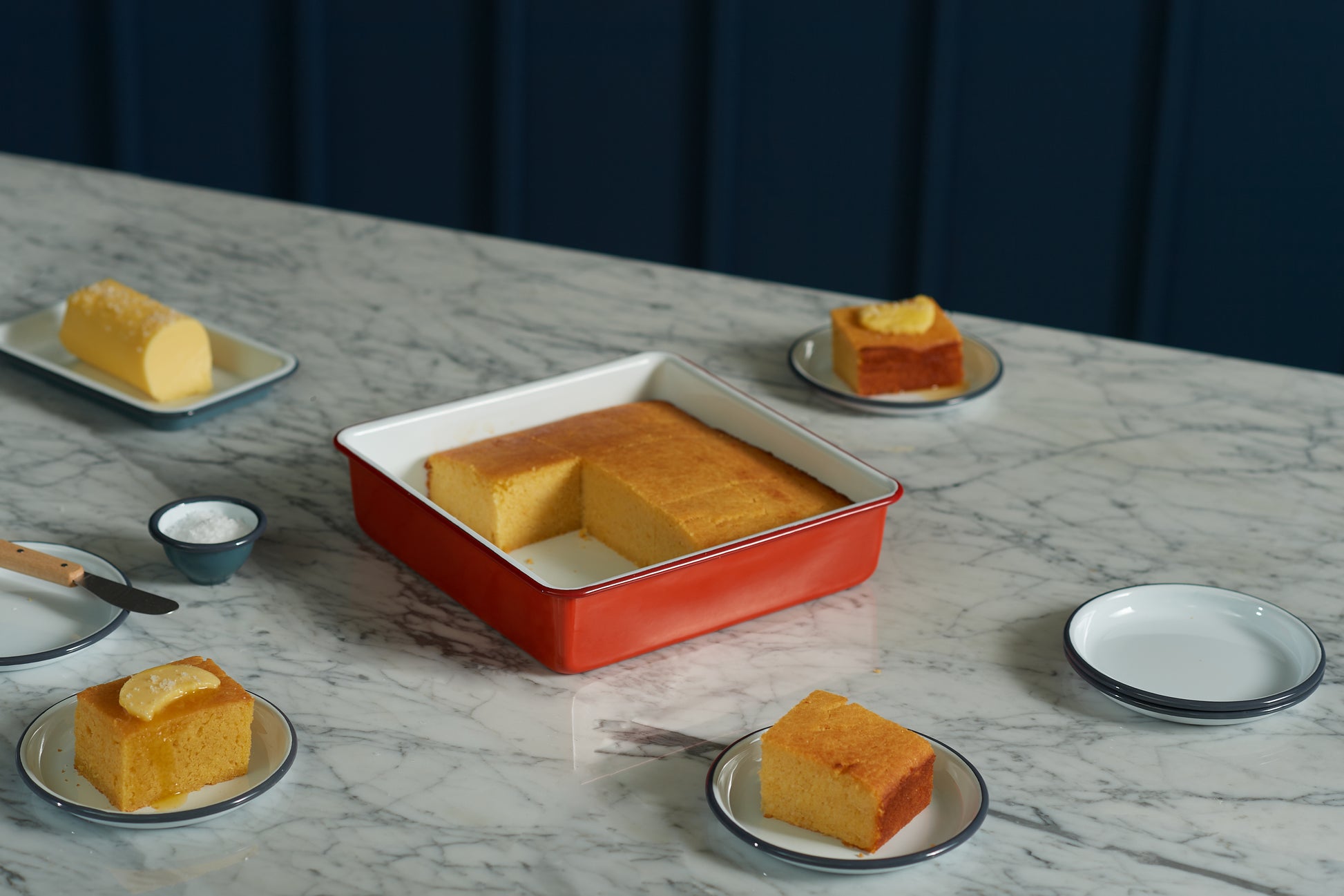 The Falcon Enamelware Square Bake Tray by Grace & Company ltd with cornbread and four plated slices on marble.