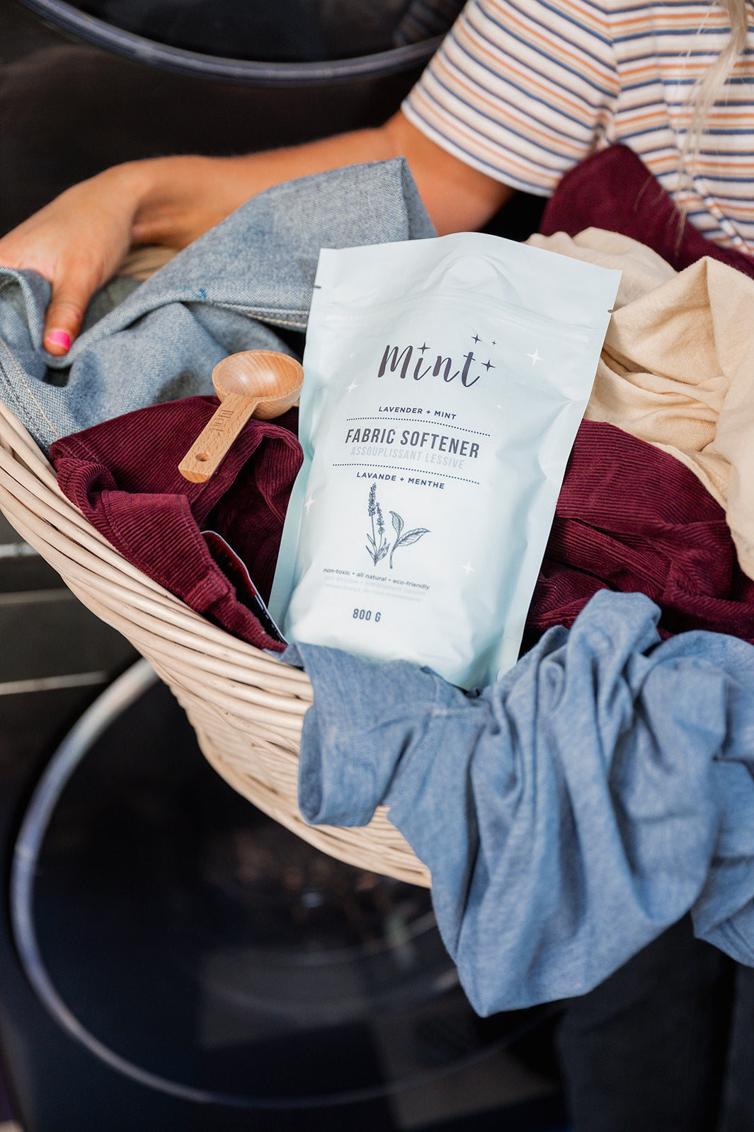 Person holding a laundry basket with clothes, a wooden scoop, and a bag of Mint fabric softener.