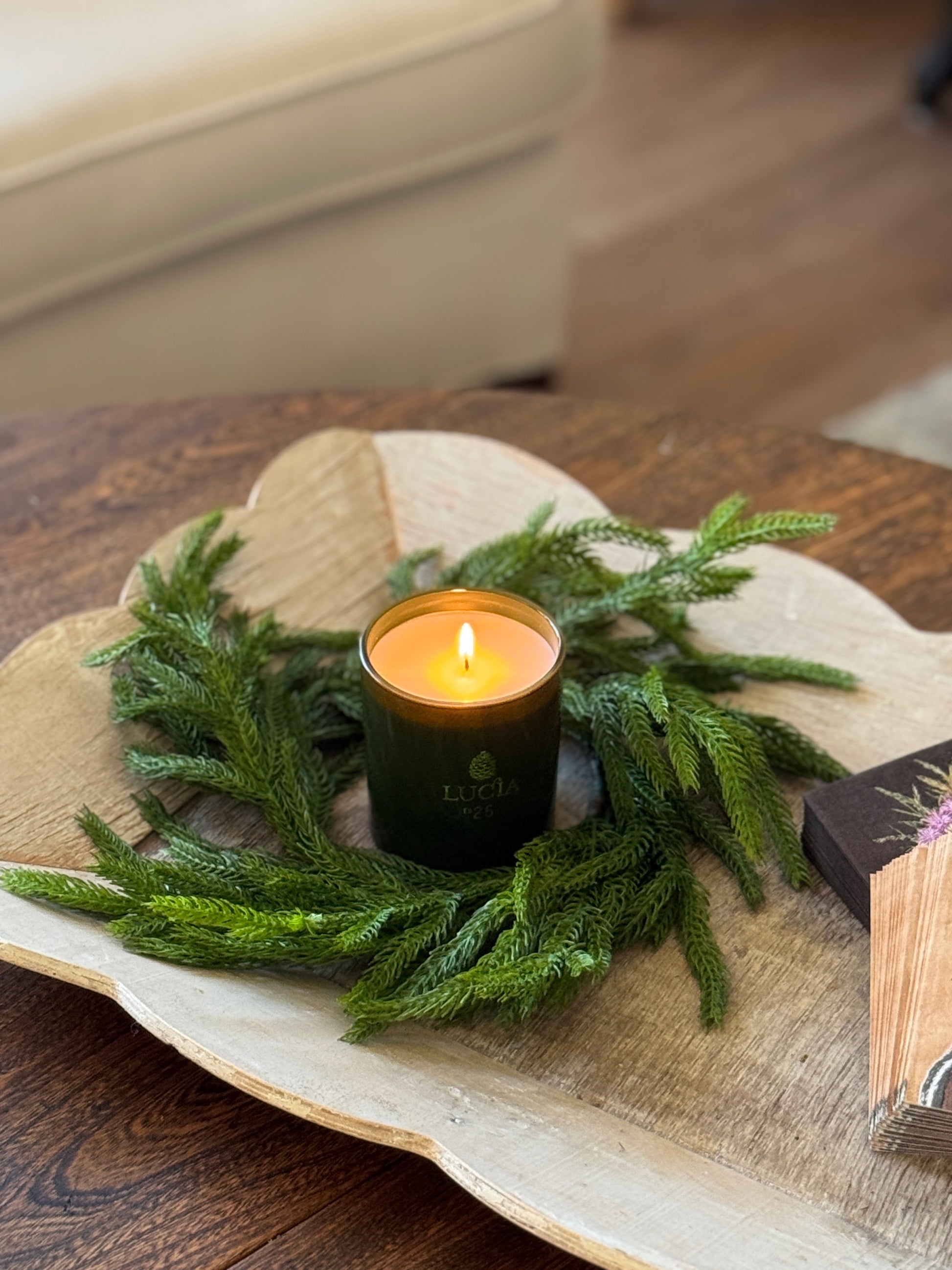 A lit green candle in a wreath of pine branches sits on a wooden tray atop a table.
