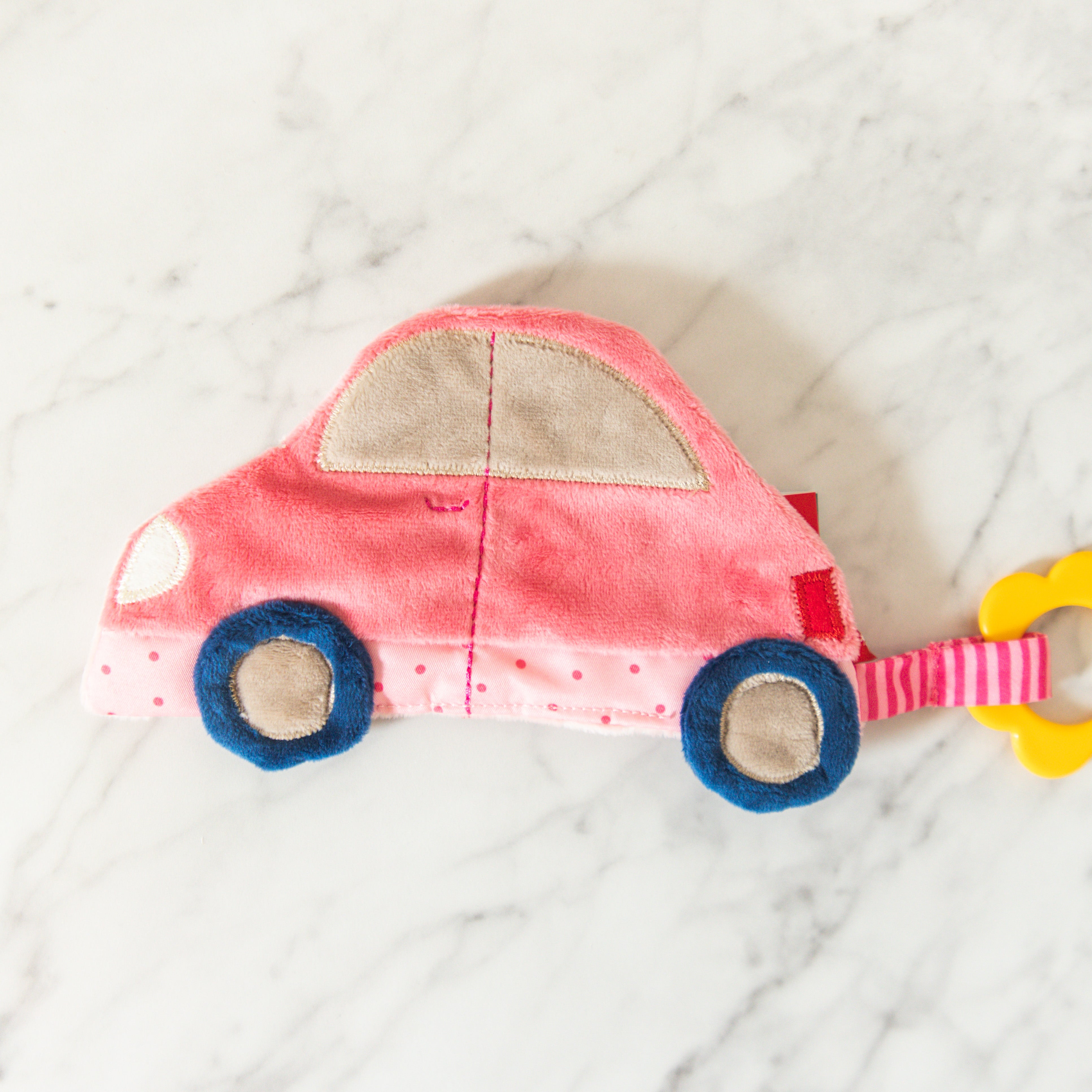 The Sigykid Crinkle Toy by Grace & Company: a pink plush car with blue wheels and yellow teether on white marble.