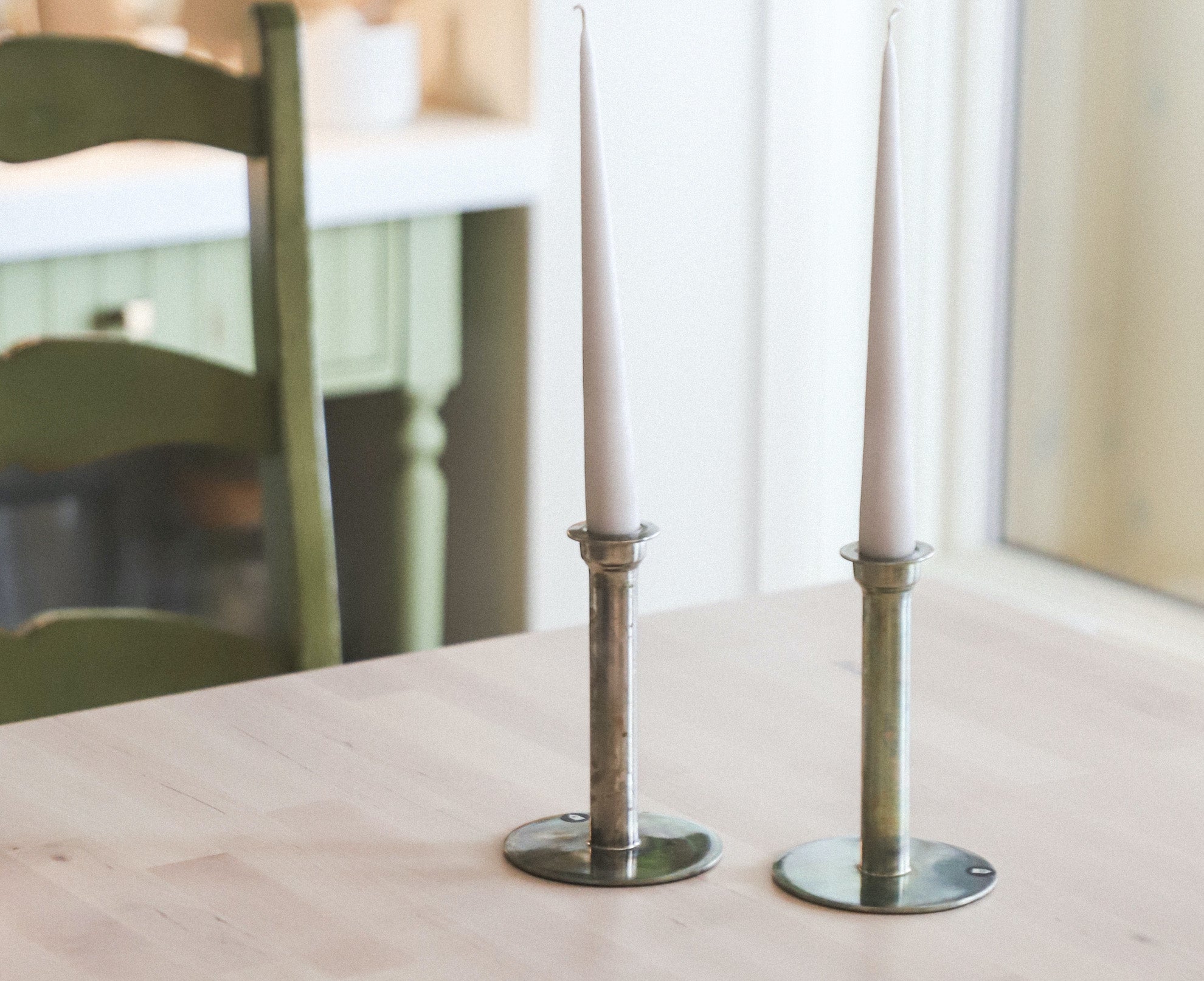 Two unlit white taper candles in metal holders on a light wooden table with a green chair in the background.
