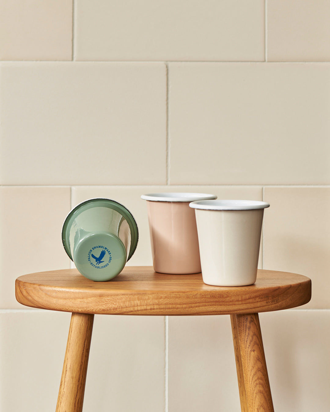 Three Grace & Company Falcon Enamelware Tumblers, one tipped over, on a wooden stool with a tiled background.