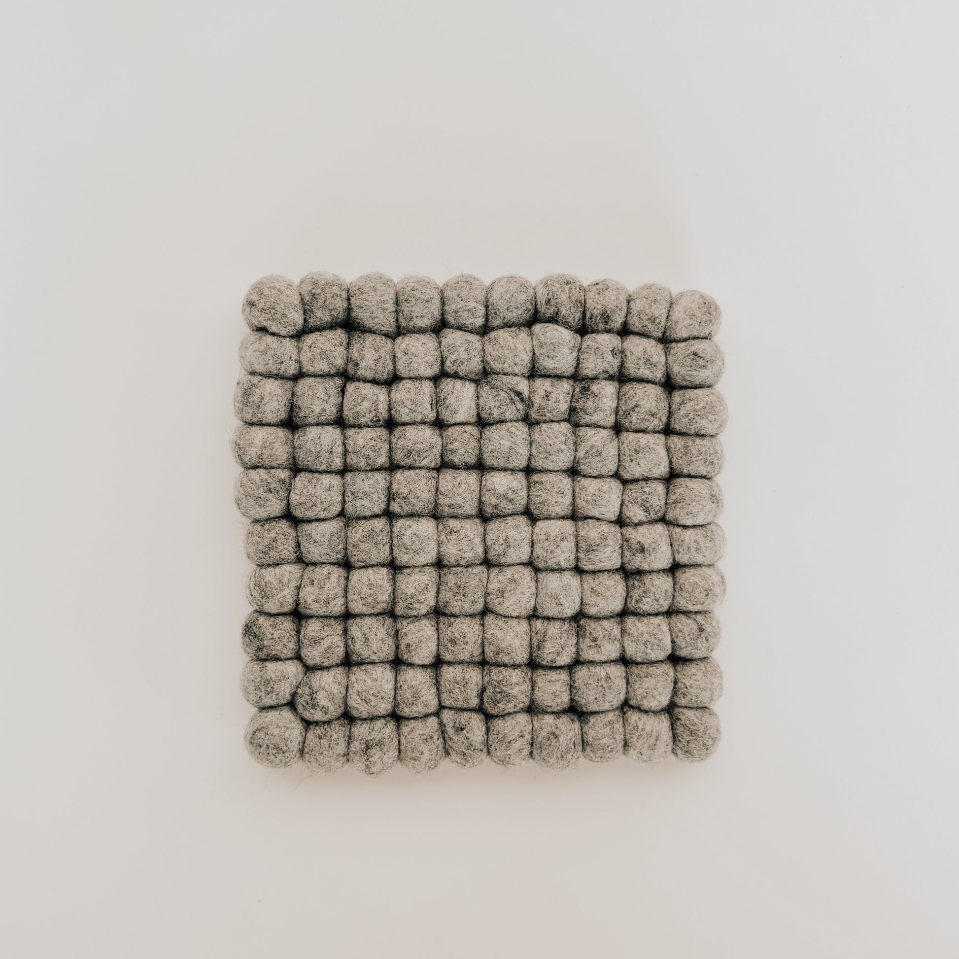 The Grace & Company Wool Felt Ball Trivet features small grey felted wool balls in a square grid on white.
