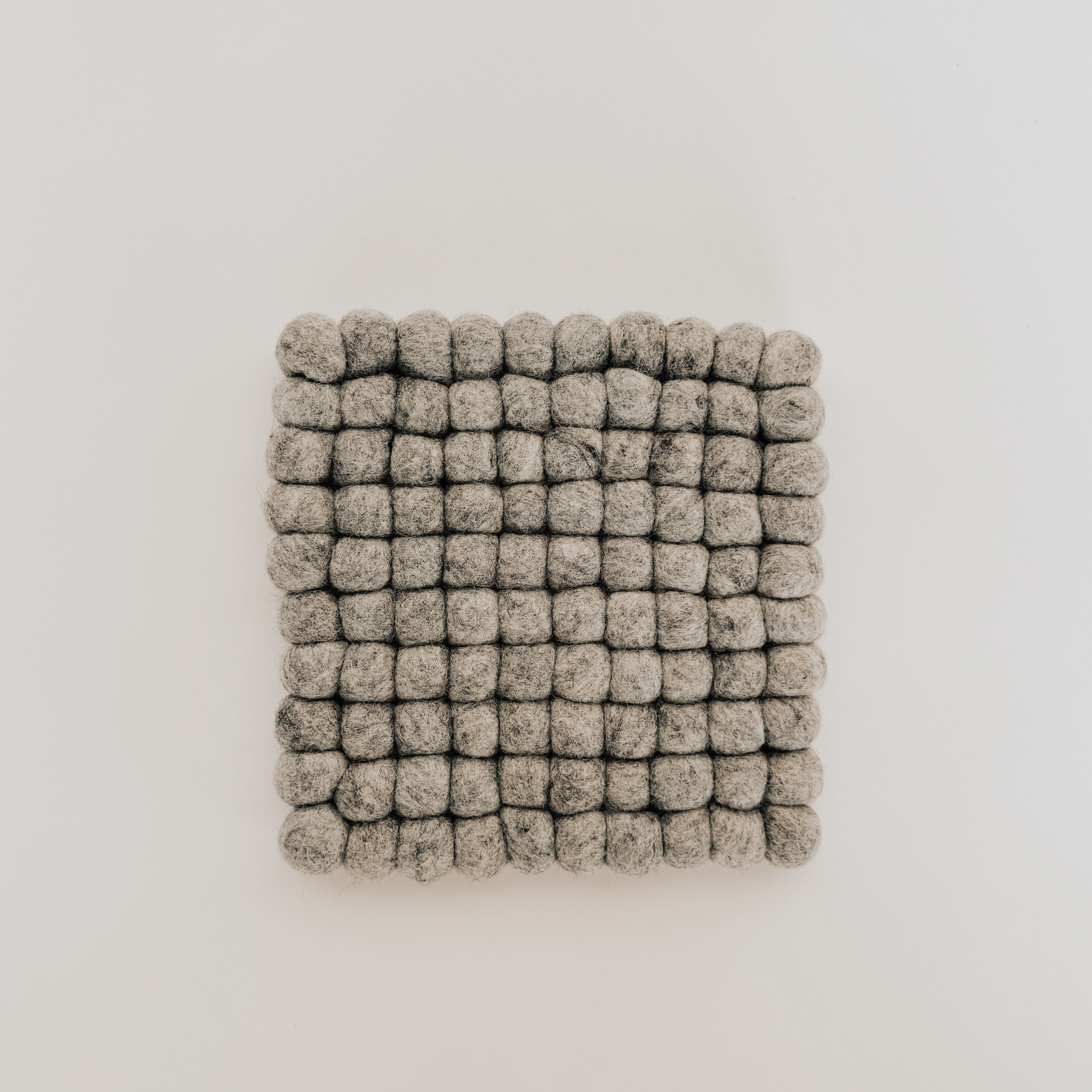 The Grace & Company Wool Felt Ball Trivet features small grey felted wool balls in a square grid on white.