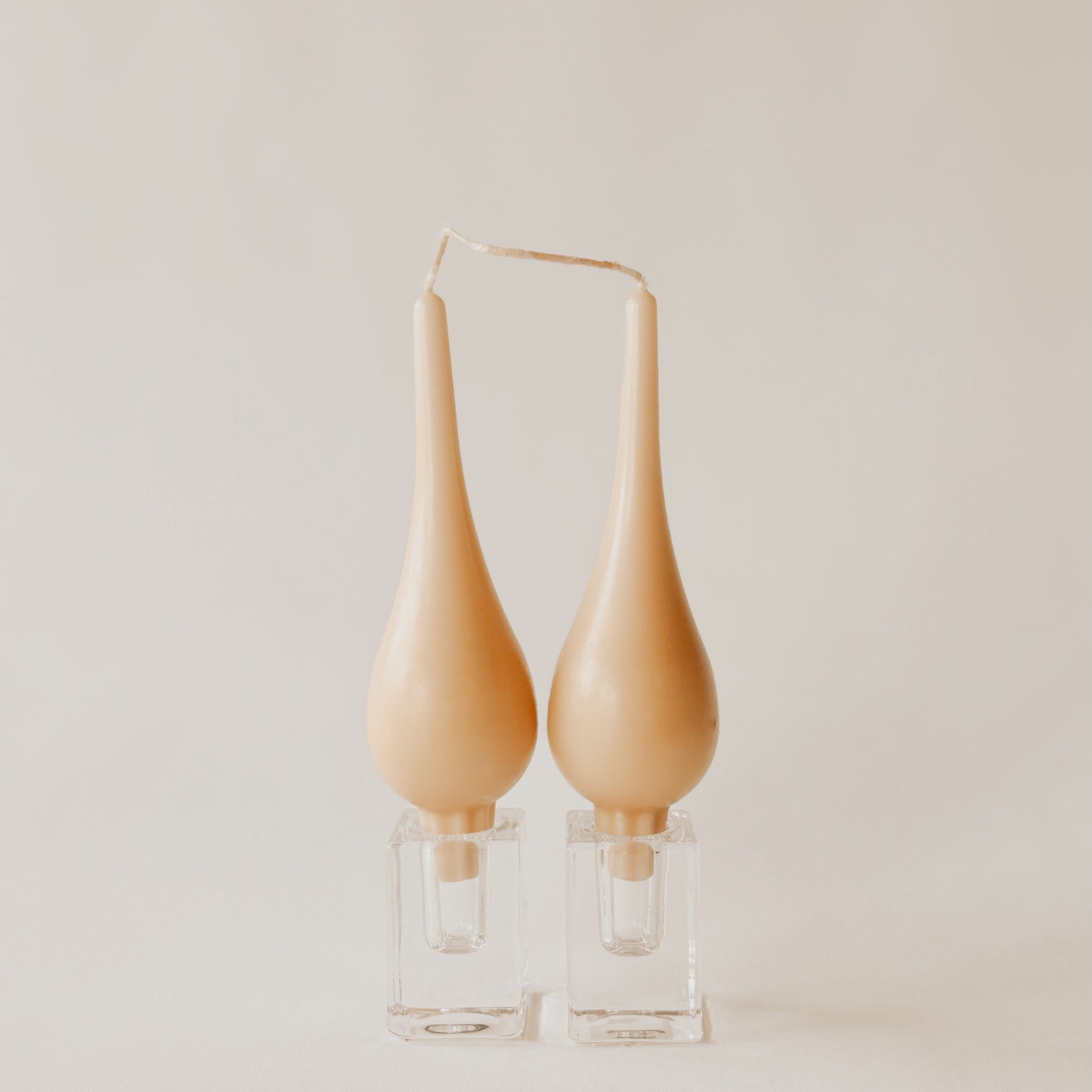 Two beige, teardrop-shaped candles joined by a single wick, standing on clear glass holders against a plain background.
