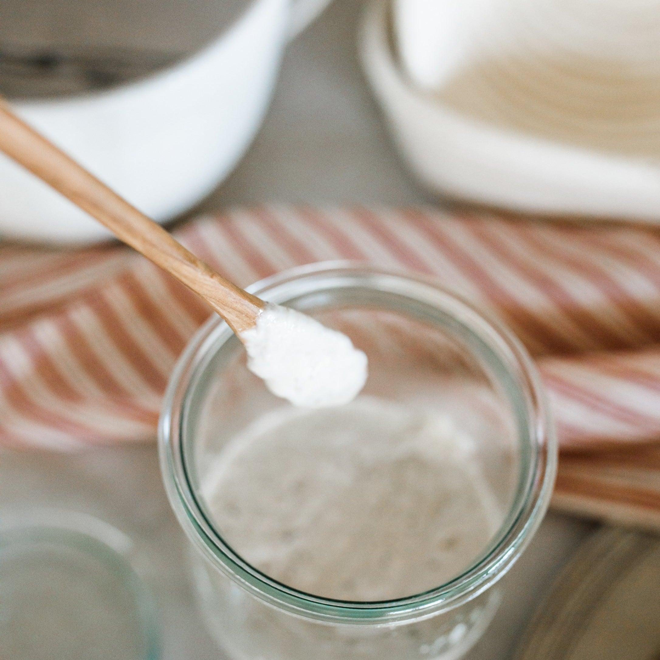 The Grace & Company ltd French Olive Wood Mustard Spoon - 7 holds sourdough starter over a glass jar with a striped cloth behind.
