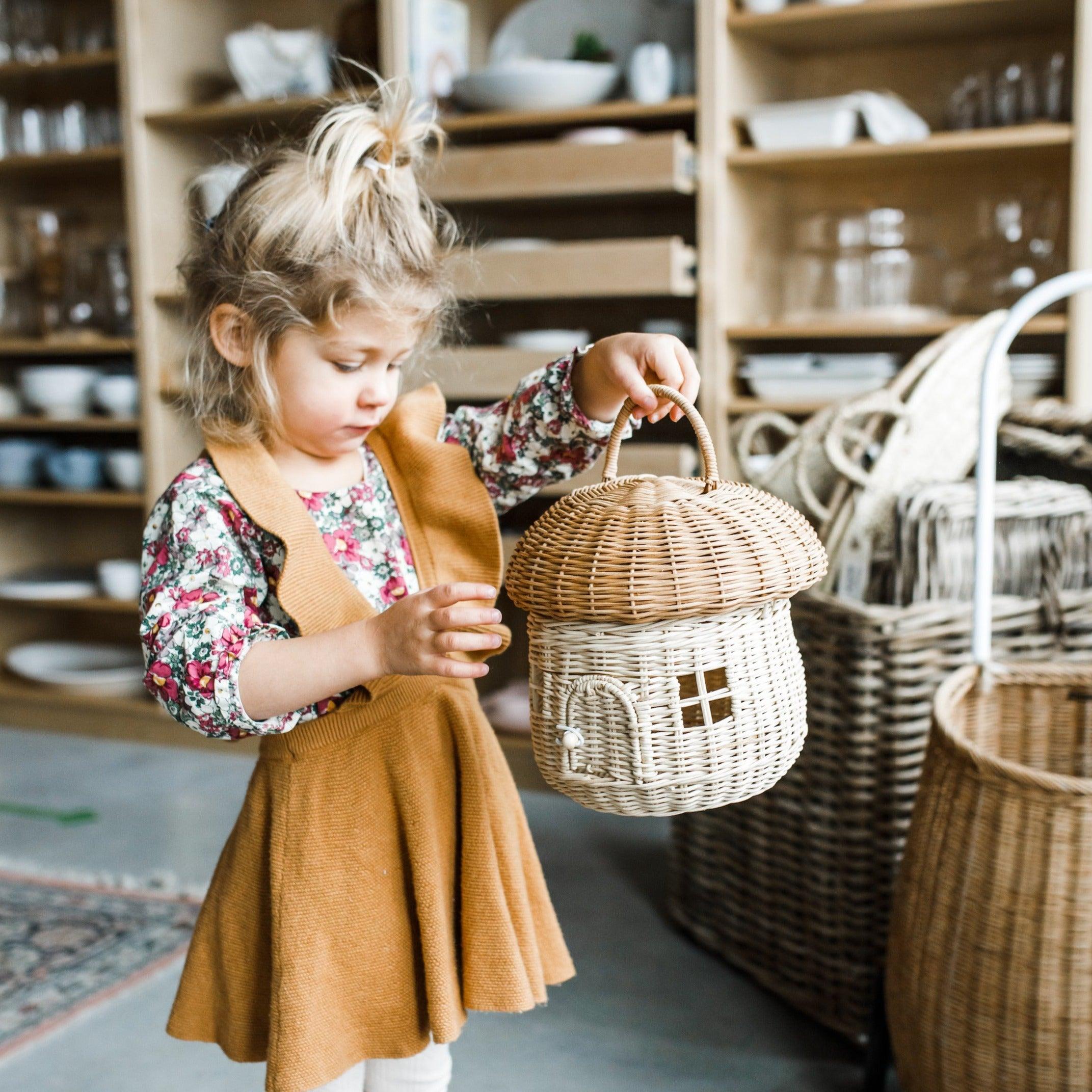 Young girl in a floral top and brown dress holds the Grace & Company Olli Ella - Mushroom Basket in a shop.