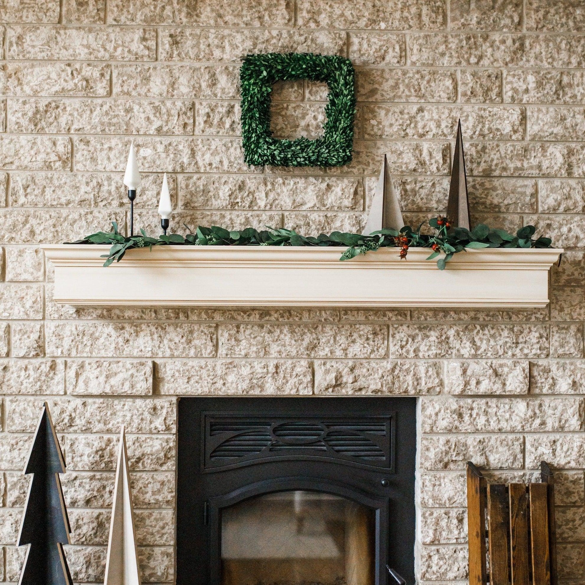 A fireplace with a white mantel, Grace & Company Eucalyptus Garland, cone trees, and a green square wreath on brick.