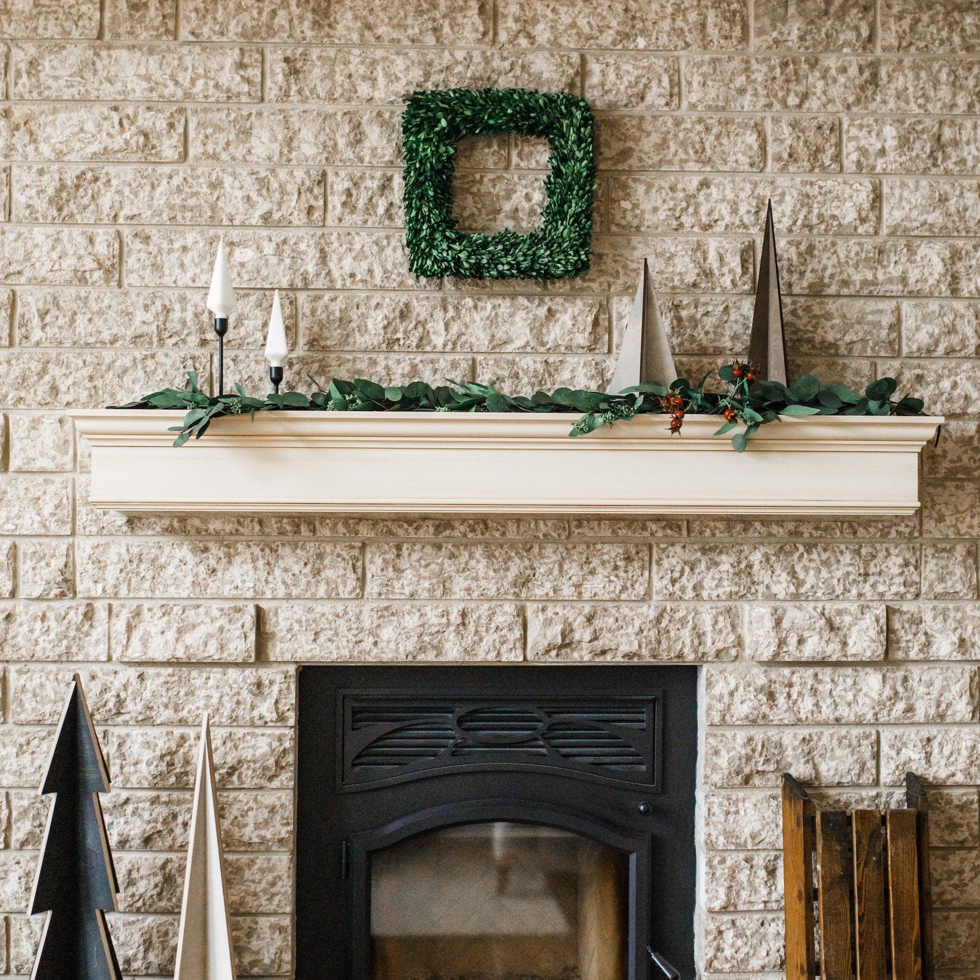 A fireplace with a white mantel, Grace & Company Eucalyptus Garland, cone trees, and a green square wreath on brick.