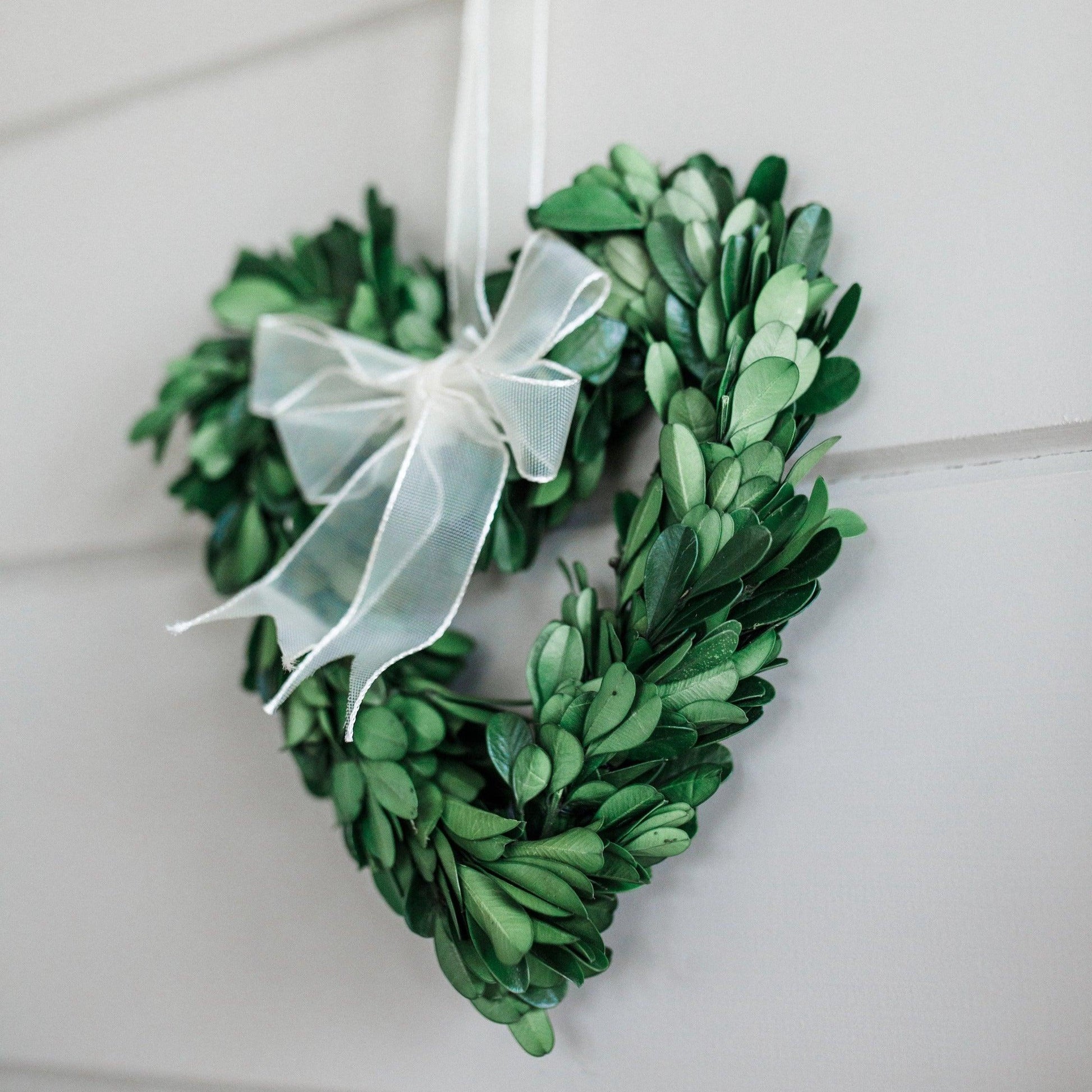 The Boxwood Heart Wreath by Grace & Company features a white ribbon bow and hangs on a white wooden wall.