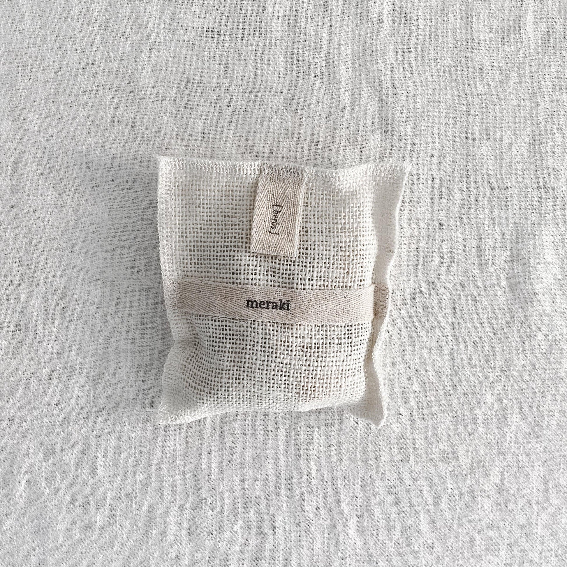 A Meraki Bath Mitt by Grace & Company on a light textured surface, featuring a small mesh pouch with label.