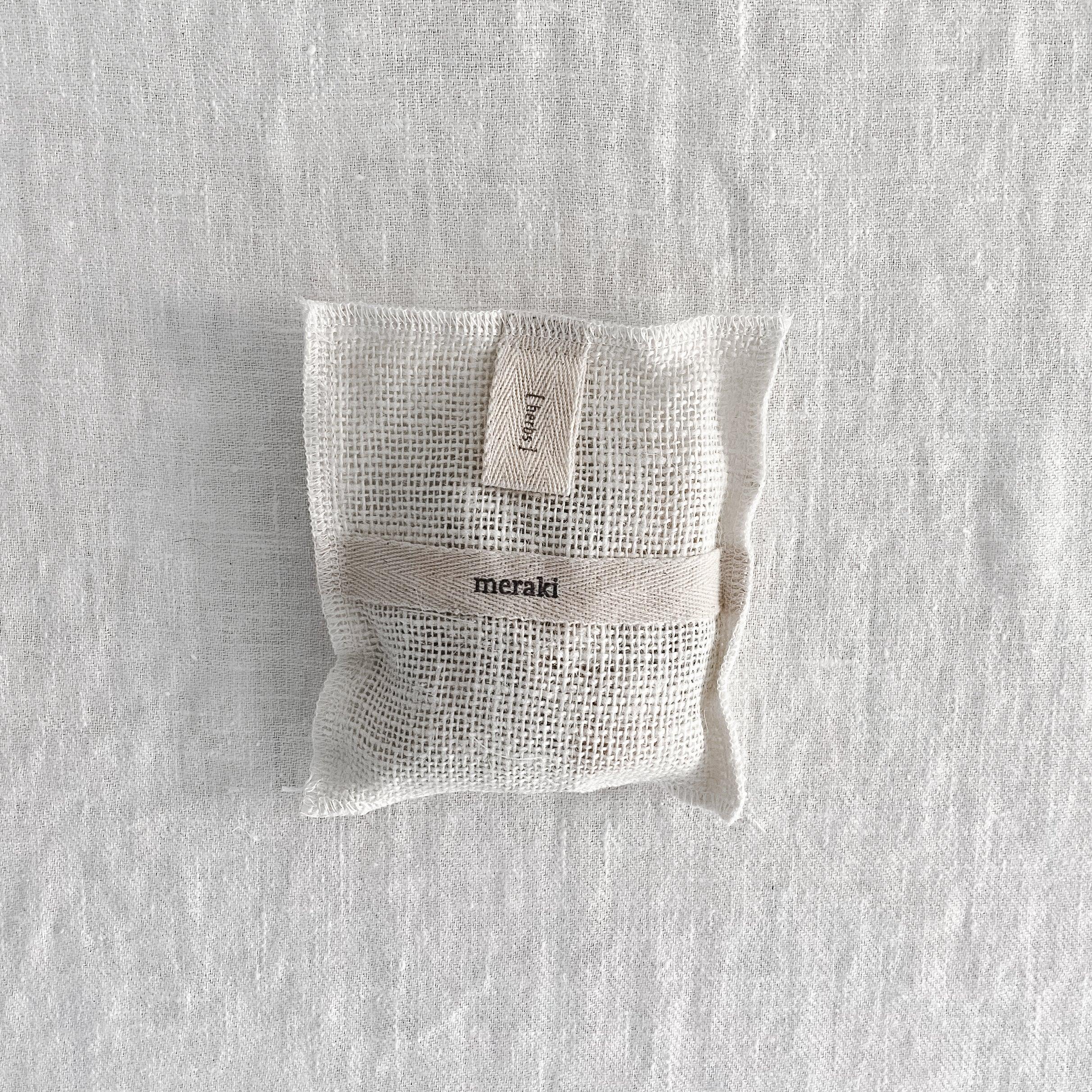 A Meraki Bath Mitt by Grace & Company on a light textured surface, featuring a small mesh pouch with label.
