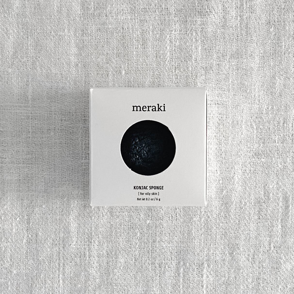 A white “Meraki - Konjac Sponge” box by Grace & Company reveals a black konjac sponge through a round window.
