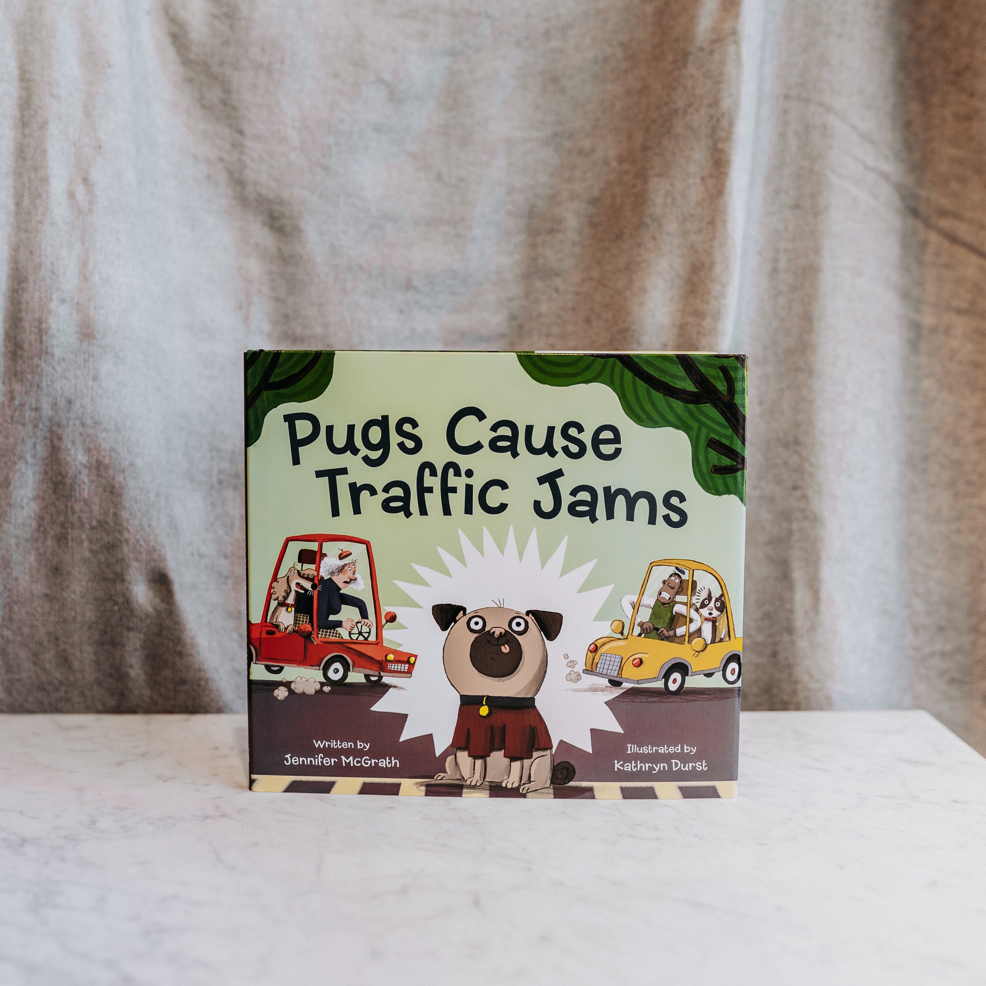 A childrens book titled Pugs Cause Traffic Jams stands on a marble surface with a soft fabric backdrop.