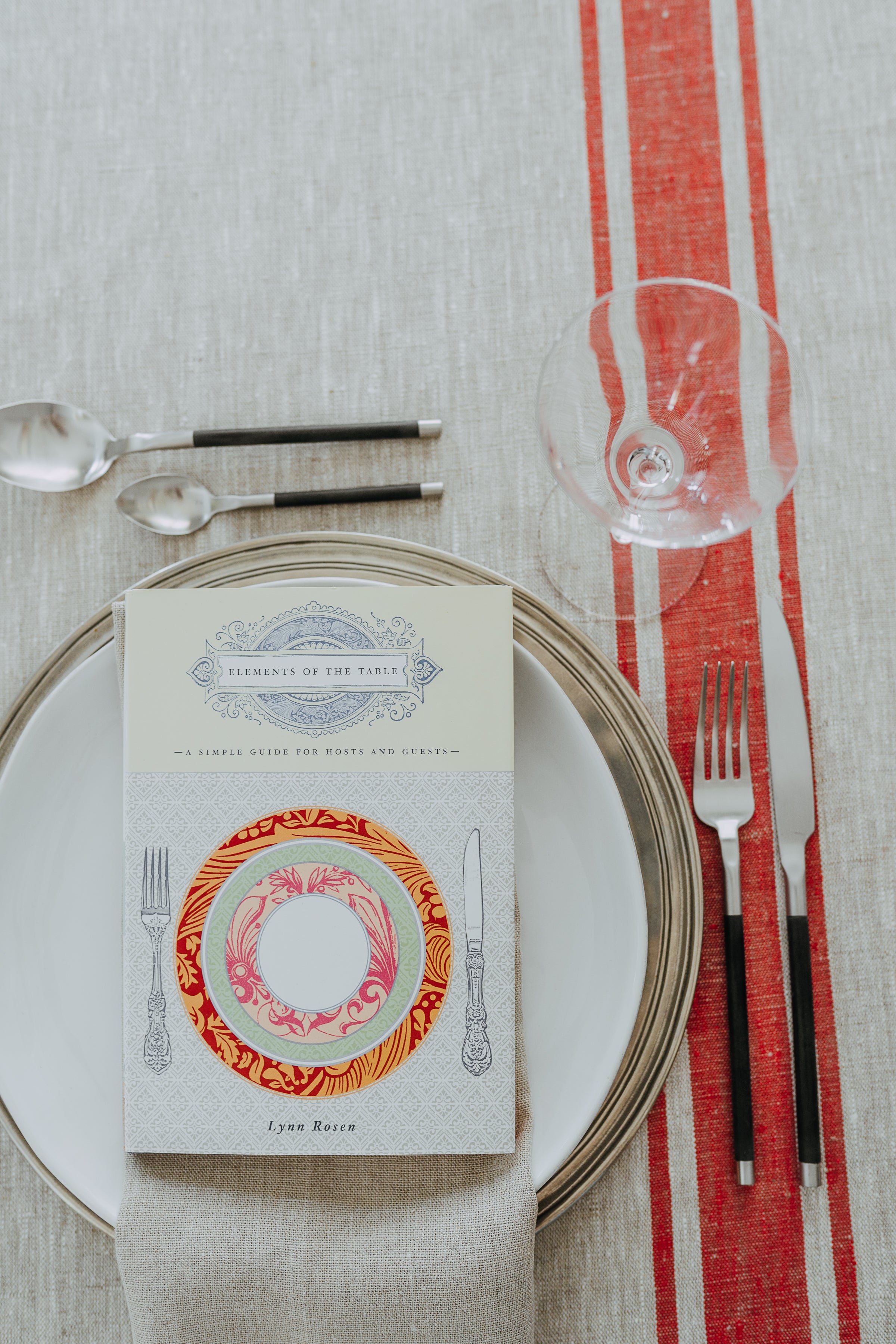 Elements of The Table by Grace & Company sits on a plate with cutlery, napkin, and empty glass on a striped tablecloth.